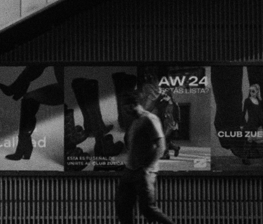 Person walking past a large advertisement poster at night, featuring various models and text including 'AW 24' and 'CLUB ZUE'.