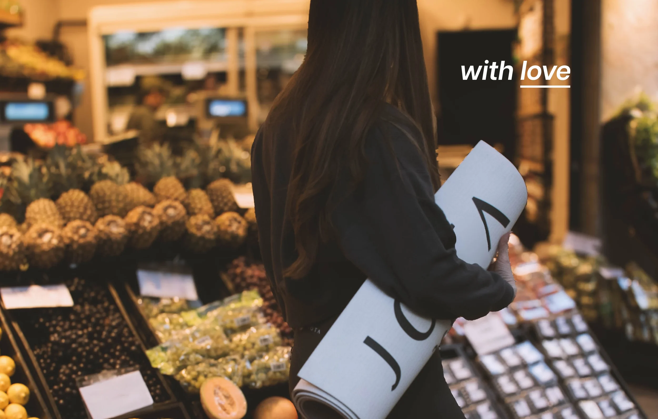 A woman in a black jacket carrying a rolled-up yoga mat while shopping for fruits at an outdoor market. The market features pineapples, grapes, and other produce displays in the background.