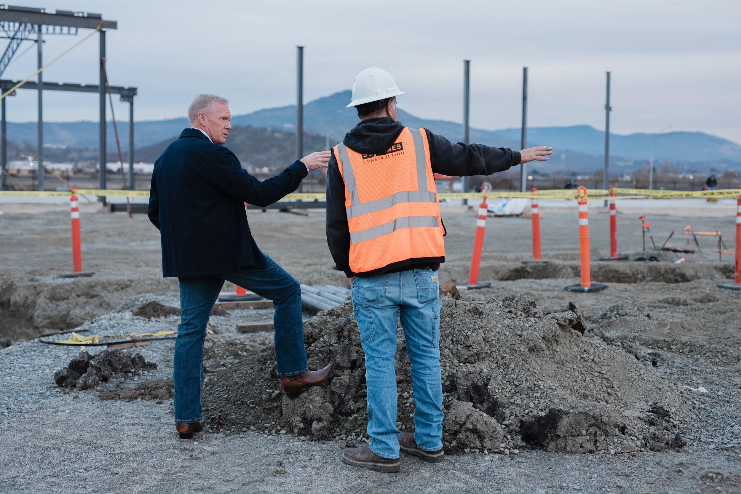 Two men at a construction site, one in a black jacket and the other in a safety vest and helmet, pointing and discussing the progress amid orange cones and caution tape.