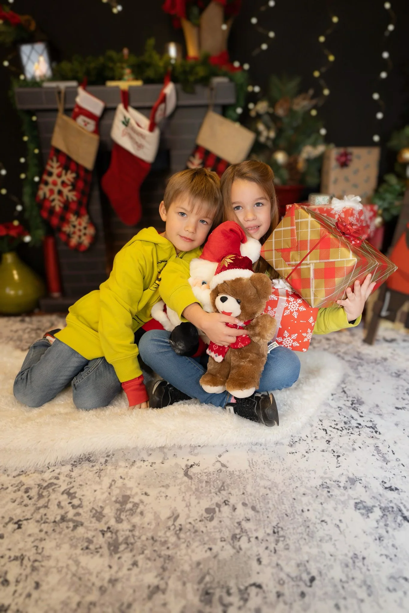 Two children sitting on a rug in front of a decorated fireplace with Christmas stockings hanging, holding presents and plush toys, celebrating Christmas.