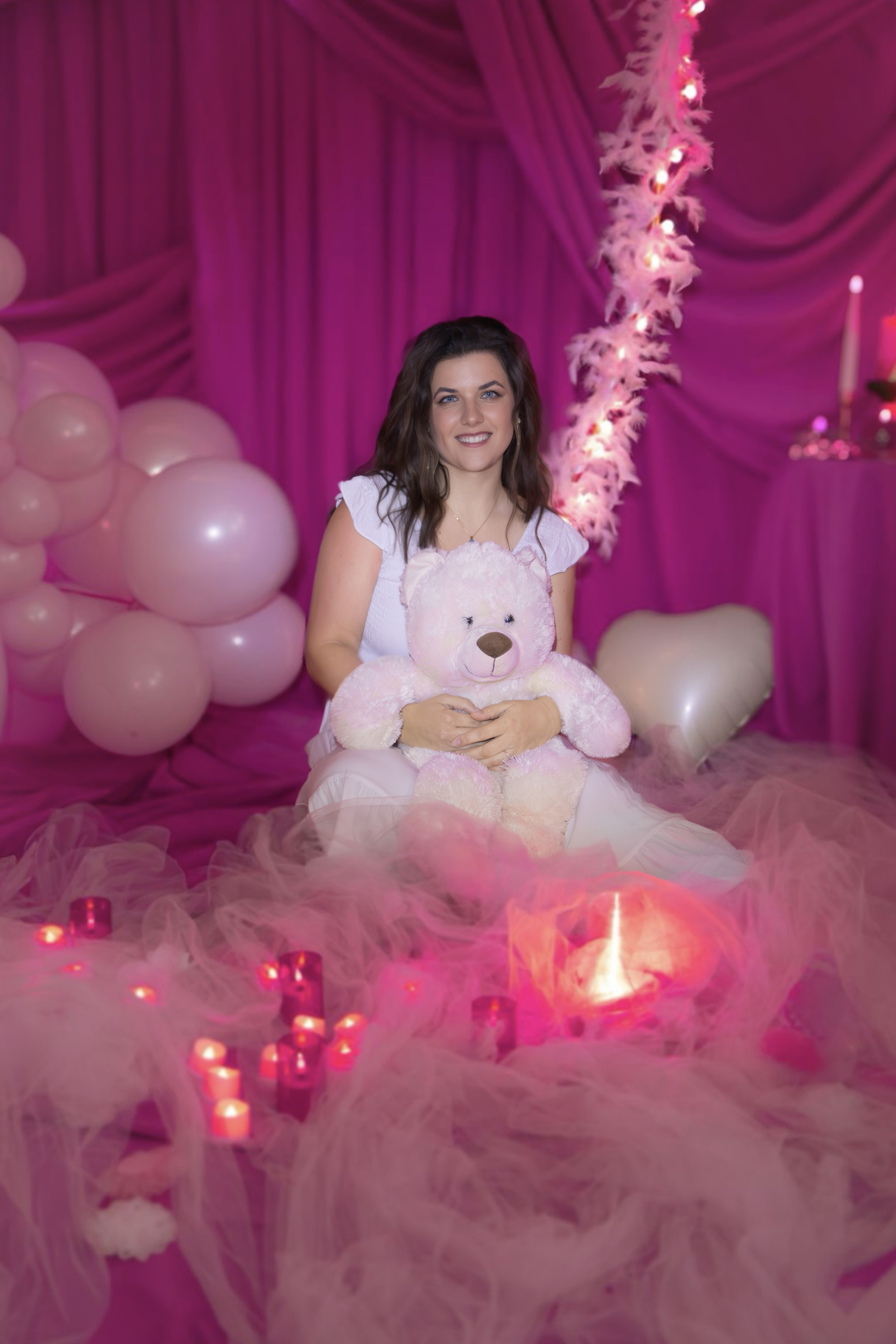 A young woman sitting on pink tulle with a large teddy bear, surrounded by pink balloons, candles, and pink curtains, in a decorated pink-themed setting with candles and soft lighting.