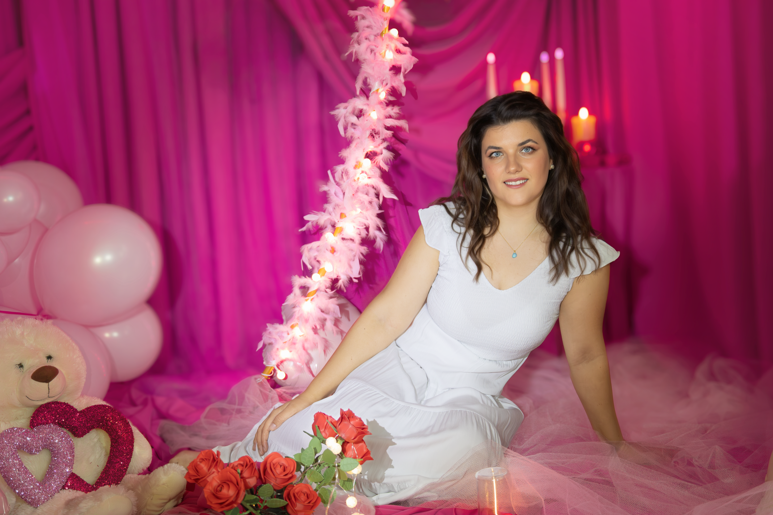 A young woman with brunette hair and blue eyes in a white dress sits on the floor in a pink-themed room with pink balloons, a pink feathered light, a pink curtain backdrop, a bouquet of red roses, a teddy bear with pink heart decorations, and lit can
