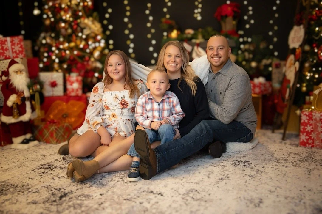 A family of four sitting on the floor in front of Christmas trees and presents, smiling for a holiday photo.