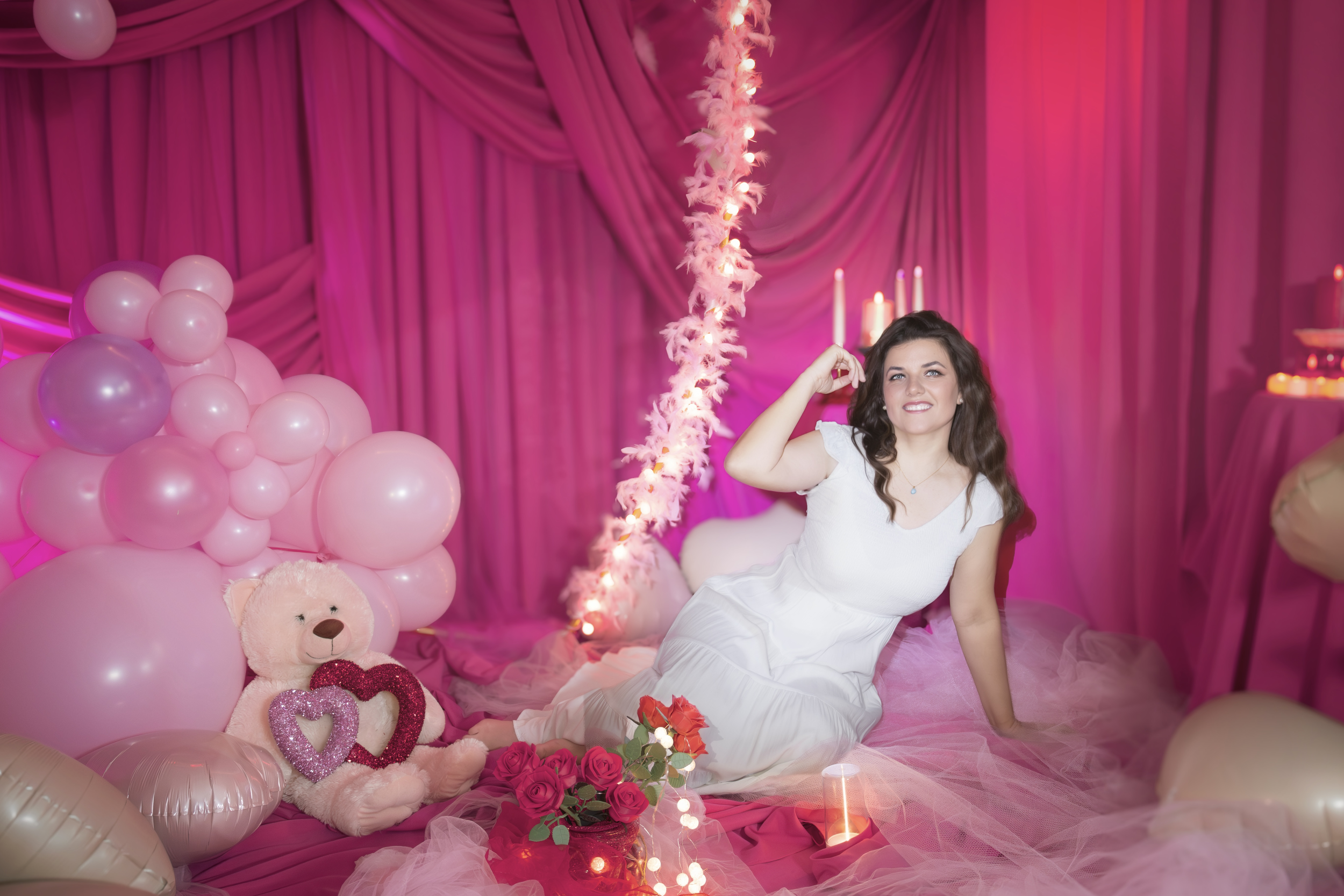A woman in a white dress sitting on a pink blanket surrounded by balloons, teddy bear, flowers, and candles with pink curtains in the background.