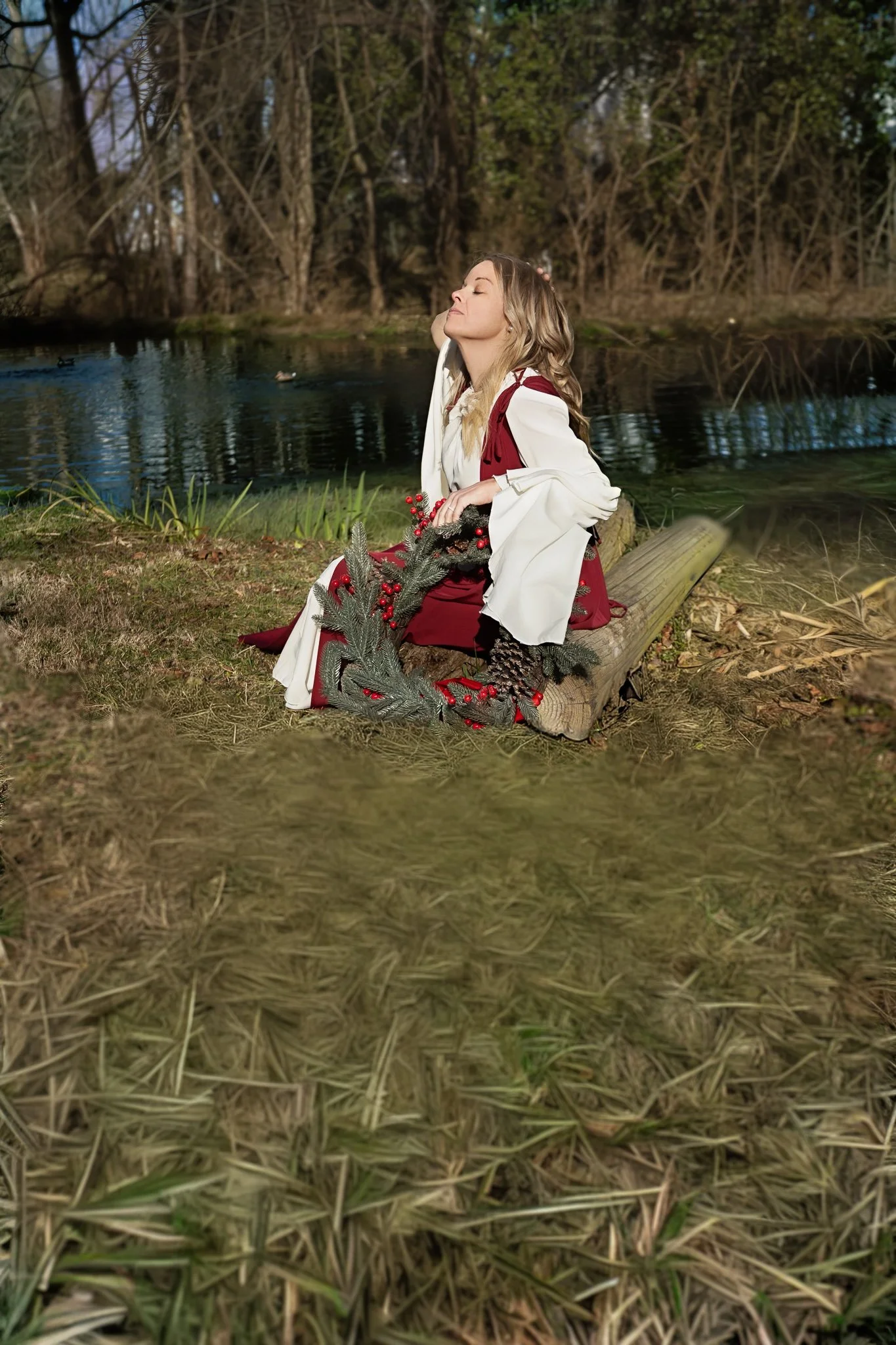 A woman with long blonde hair sitting on a log near a pond, eyes closed, enjoying the outdoor scenery during autumn. She is dressed in a white and maroon outfit and holding holiday decorations including pine cones and evergreen branches.