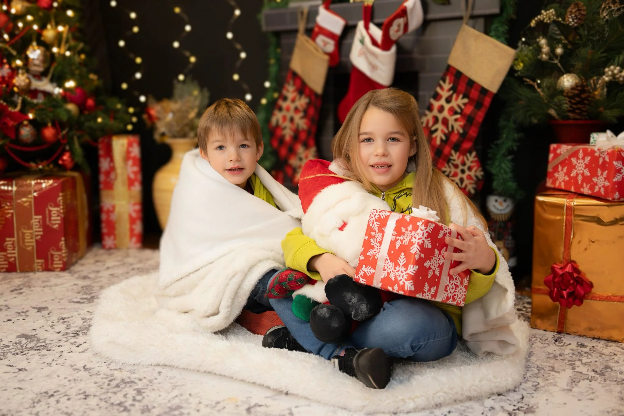 Two children sitting on a fluffy rug in front of Christmas decorated fireplace. The girl is holding a gift and a plush Santa, and the boy is wrapped in a white blanket. Christmas stockings and presents are visible in the background.