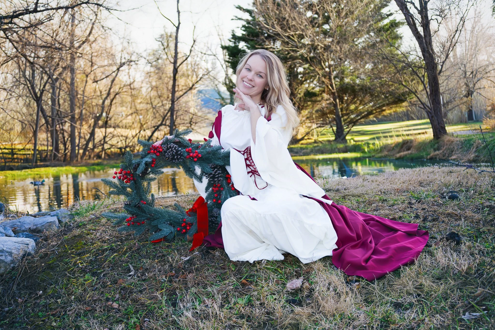 Woman in a white and burgundy dress sitting outdoors near a pond, holding a Christmas wreath, with leafless trees and a grassy area in the background.