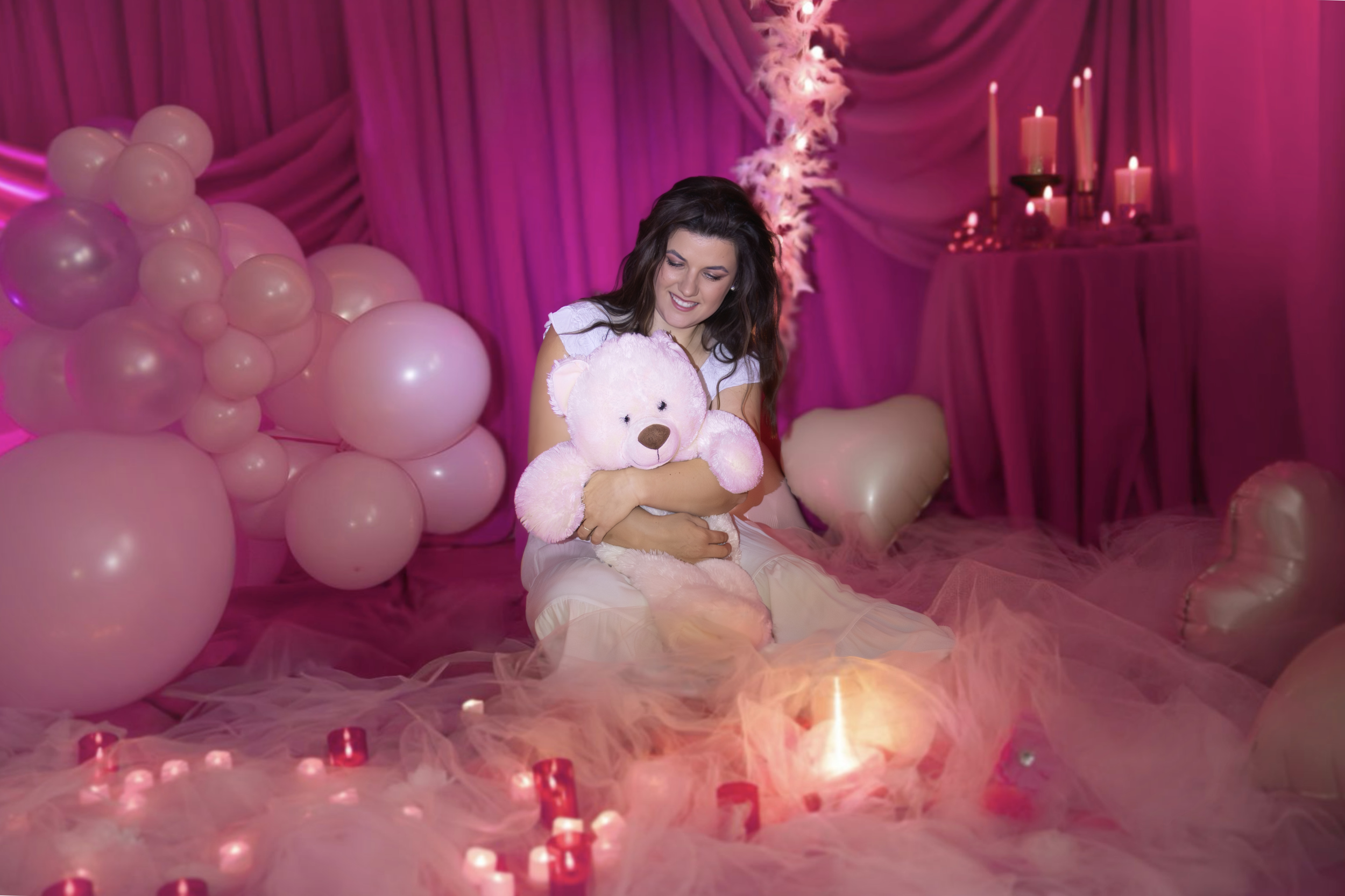 Woman in white dress sitting on pink tulle fabric, holding a pink teddy bear, in a room decorated with pink balloons, candles, and pink drapes, creating a romantic and cozy atmosphere.