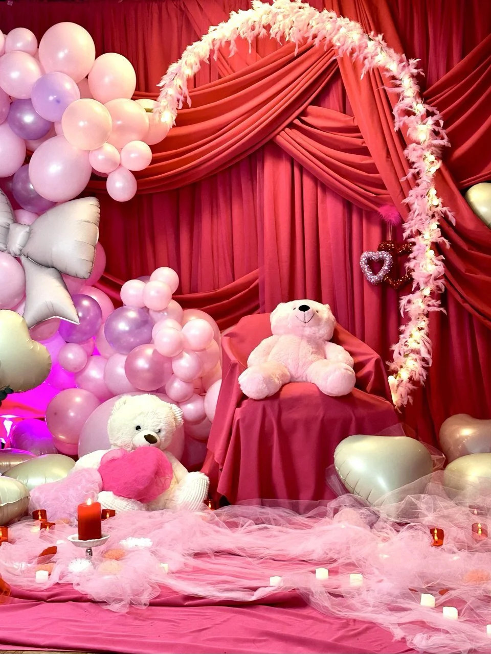 A decorated romantic or Valentine's Day scene with pink and purple balloons, teddy bears, candles, and red drapes with a feathered heart-shaped garland.