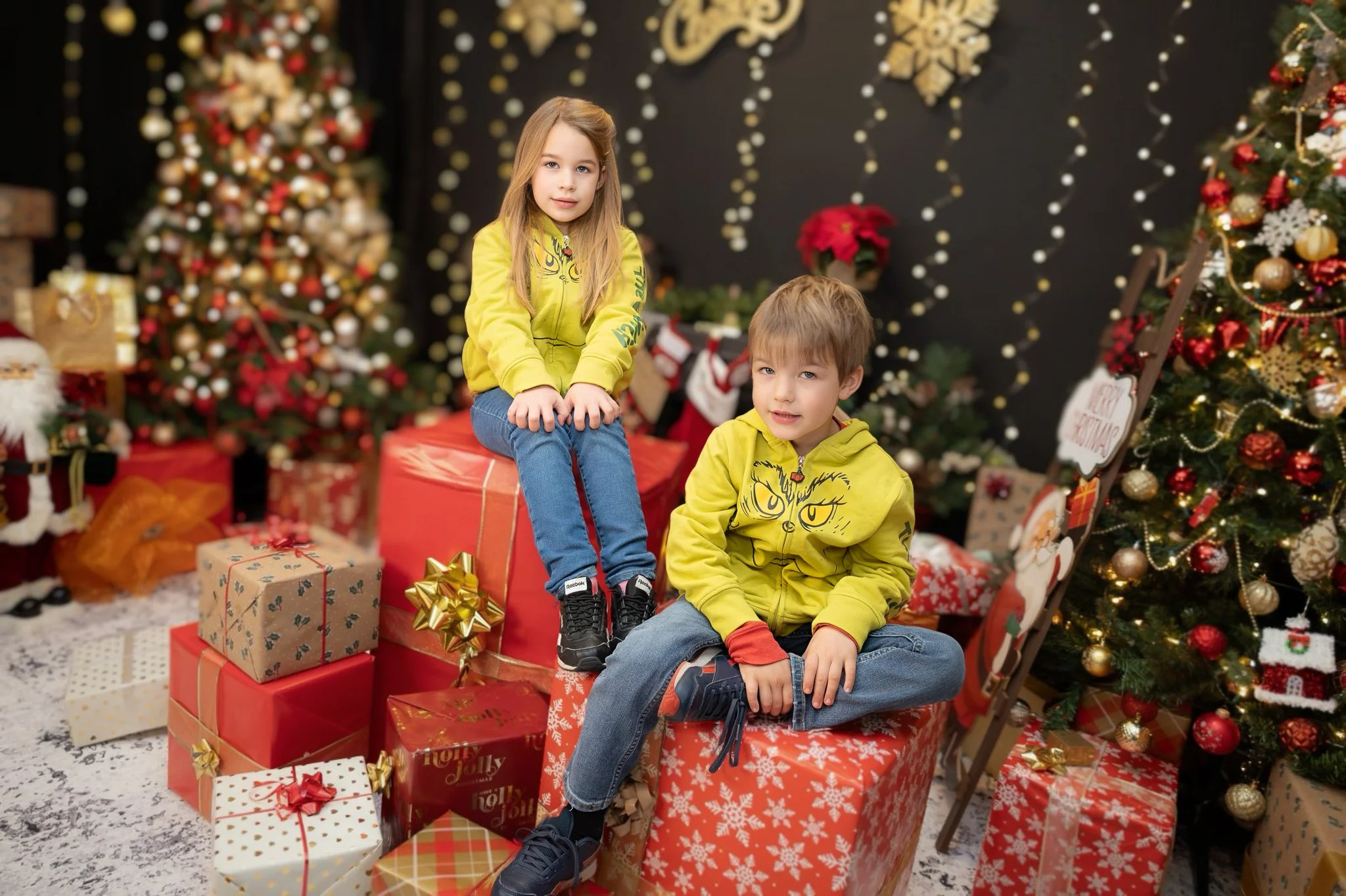 Two children, a girl and a boy, sitting on a large wrapped Christmas present in front of decorated Christmas trees, surrounded by wrapped gifts, with Christmas decorations and a black wall with lights and snowflake decorations in the background.
