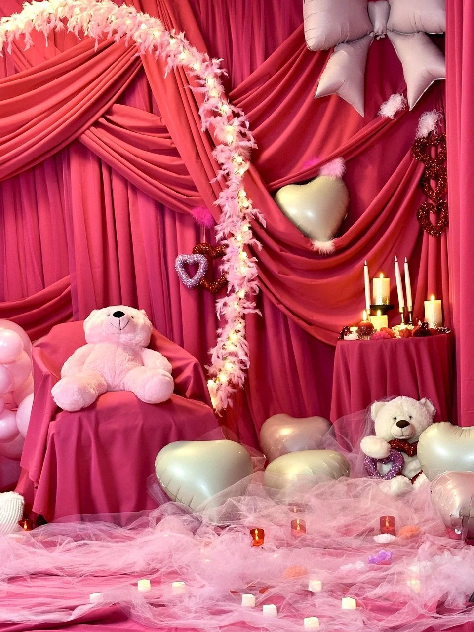 A pink-themed decorative setup with teddy bears, heart-shaped balloons, candles, and pink drapery, creating a romantic or Valentine's Day ambiance.