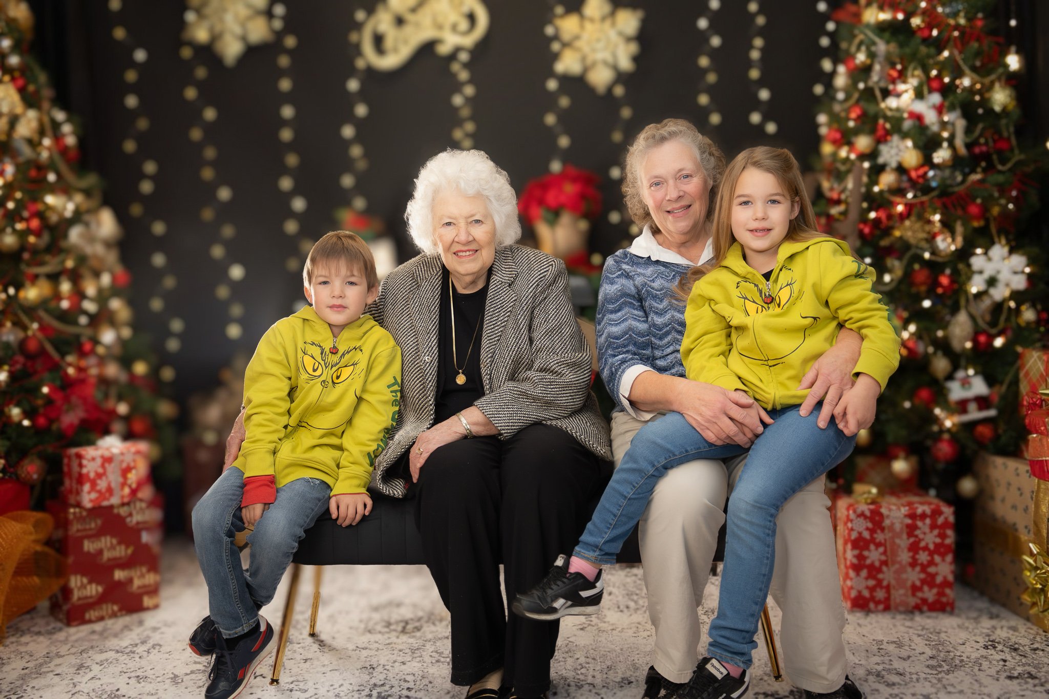 A multigenerational family of five celebrating Christmas indoors, sitting in front of decorated Christmas trees with presents around them.