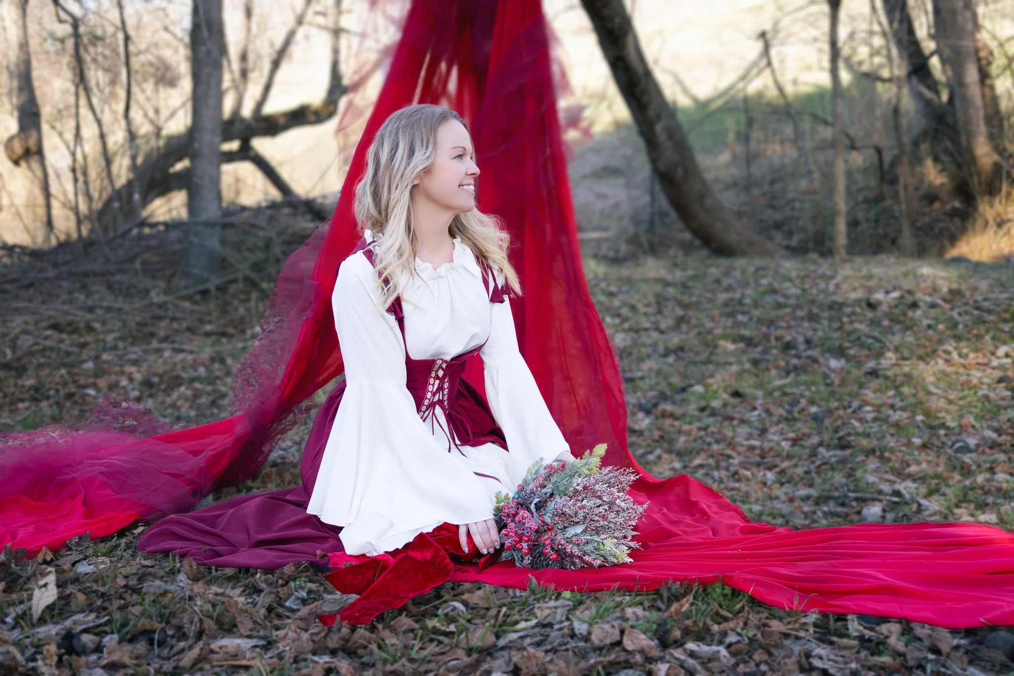 A woman in historical costume sitting on the ground with a bouquet of flowers, outside in a wooded area, with a red sheer fabric draped behind her.