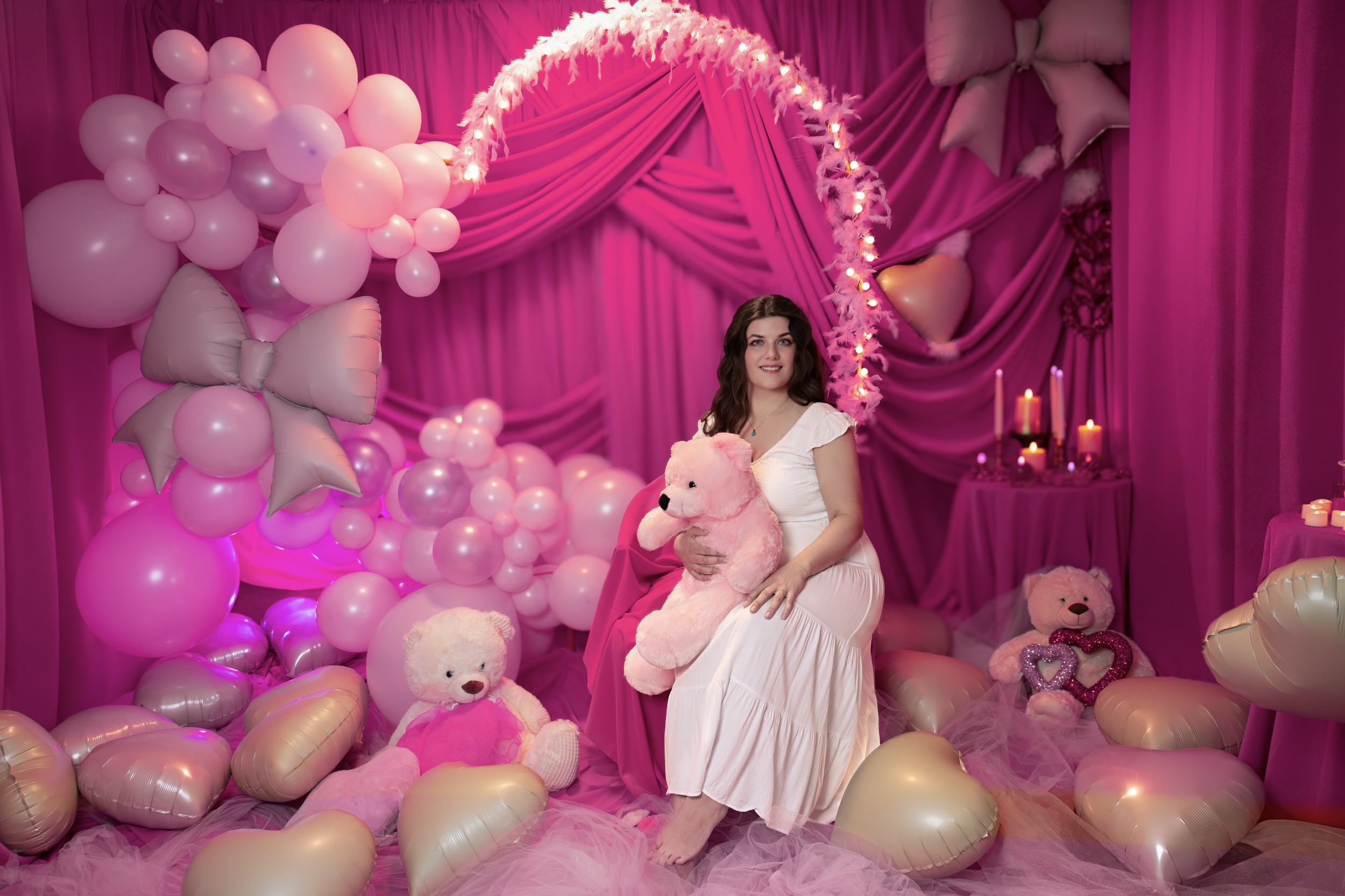 Young woman in a white dress sitting on a pink draped stage decorated with pink balloon arrangements, holding a pink teddy bear, surrounded by more teddy bears, pink hearts, and candles, with pink curtains in the background.