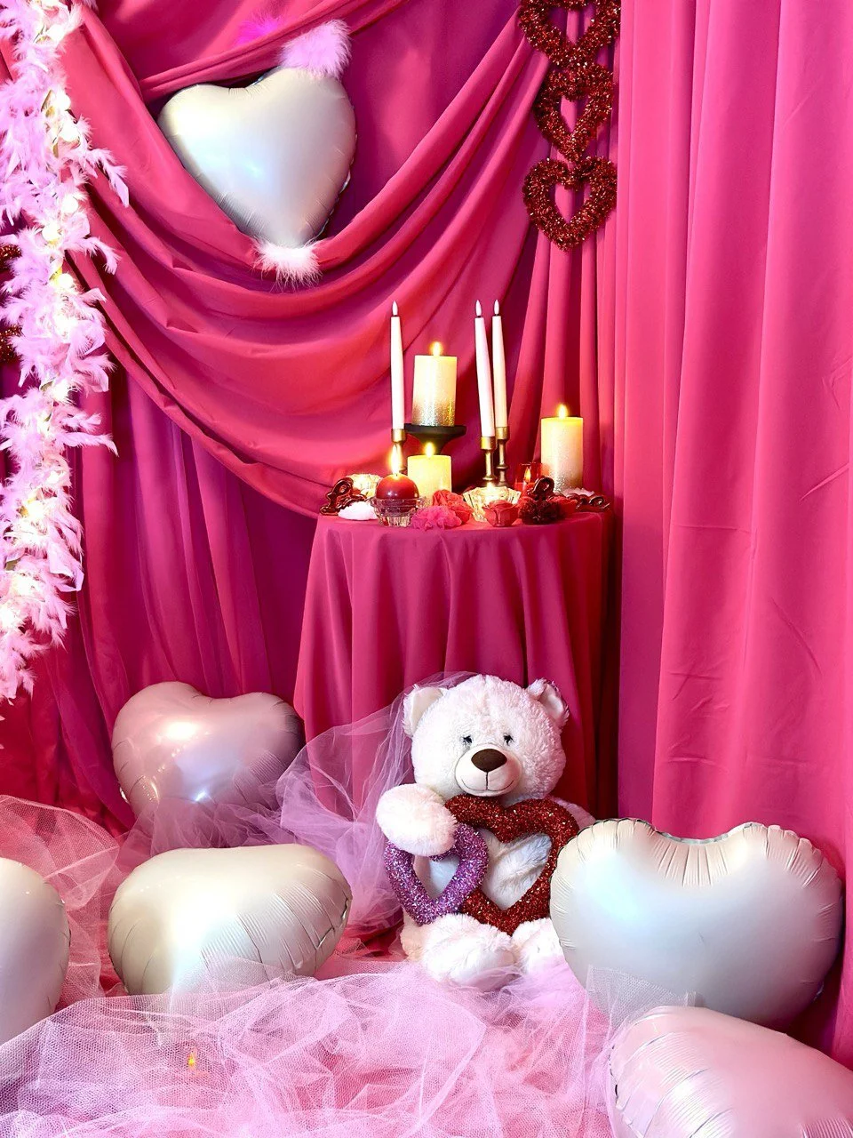 A Valentine-themed display with pink drapery, heart-shaped balloons, lit candles, a white teddy bear wearing a veil, and heart-shaped decorations.