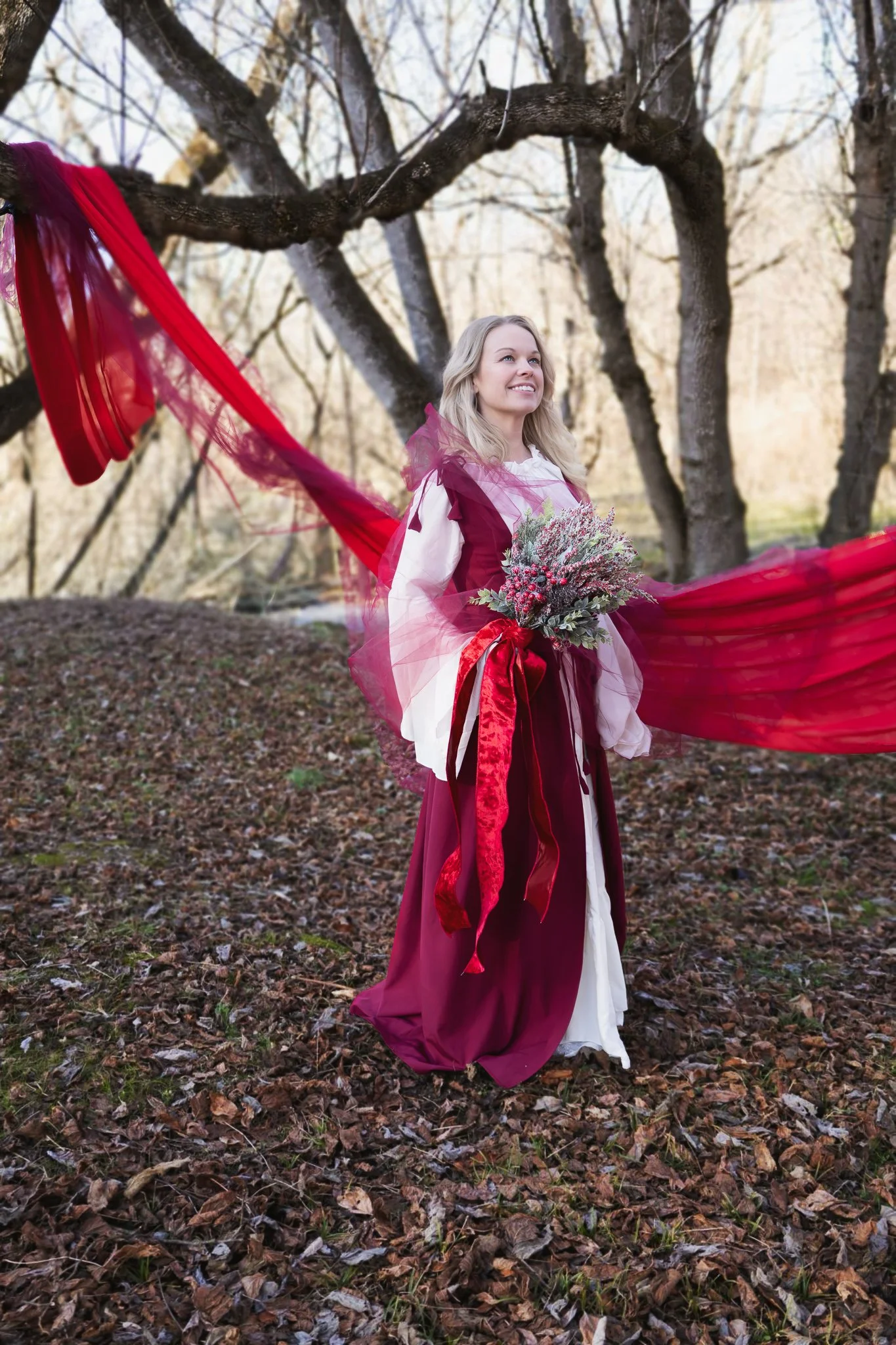 A smiling woman in a dress holding a bouquet of flowers standing outdoors with red fabric or ribbons flowing around her and bare trees in the background.