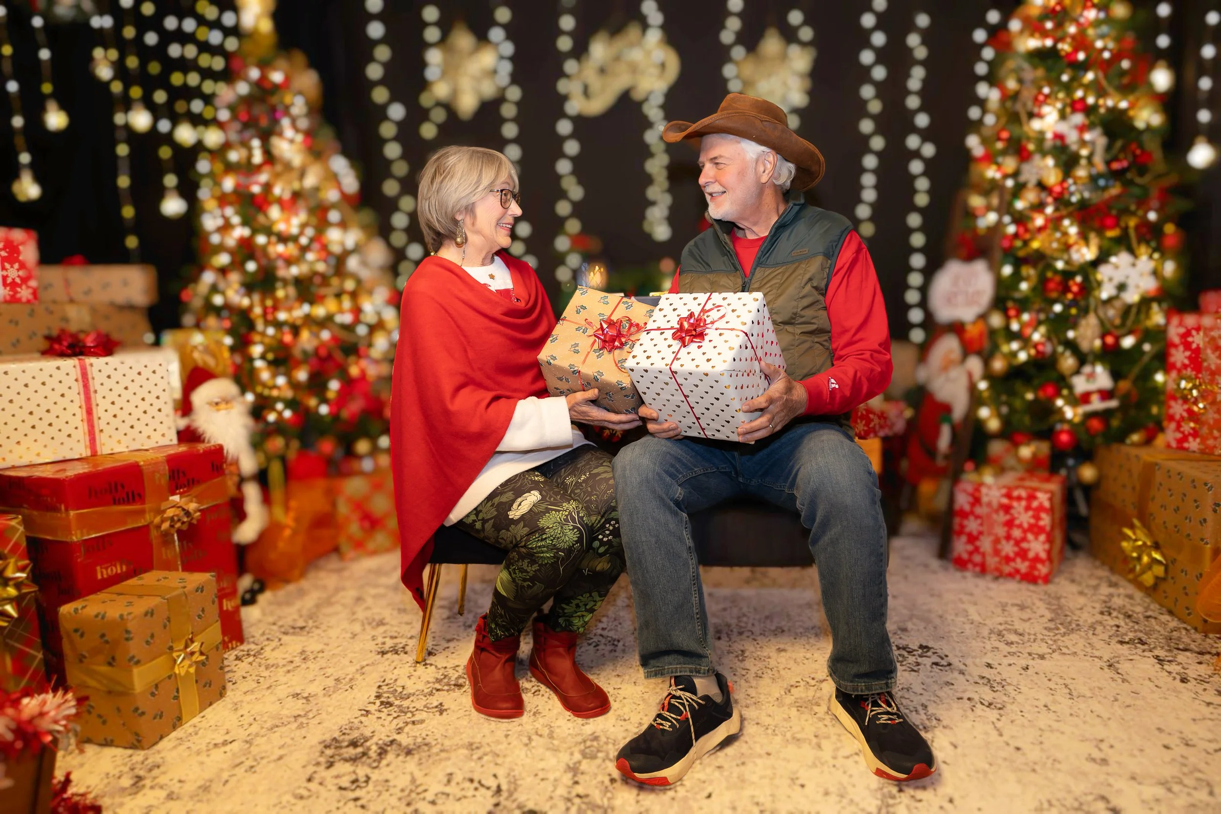 An elderly woman and man exchanging Christmas presents in front of decorated Christmas trees, with wrapped gifts around them, in a festive holiday setting.