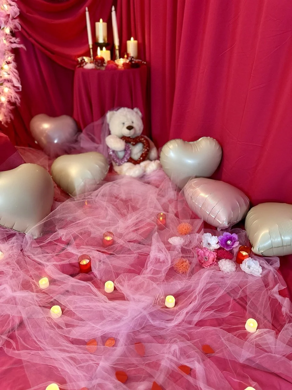 Pink-themed display with candles, heart-shaped balloons, a teddy bear wearing a veil, flowers, and pink tulle fabric.