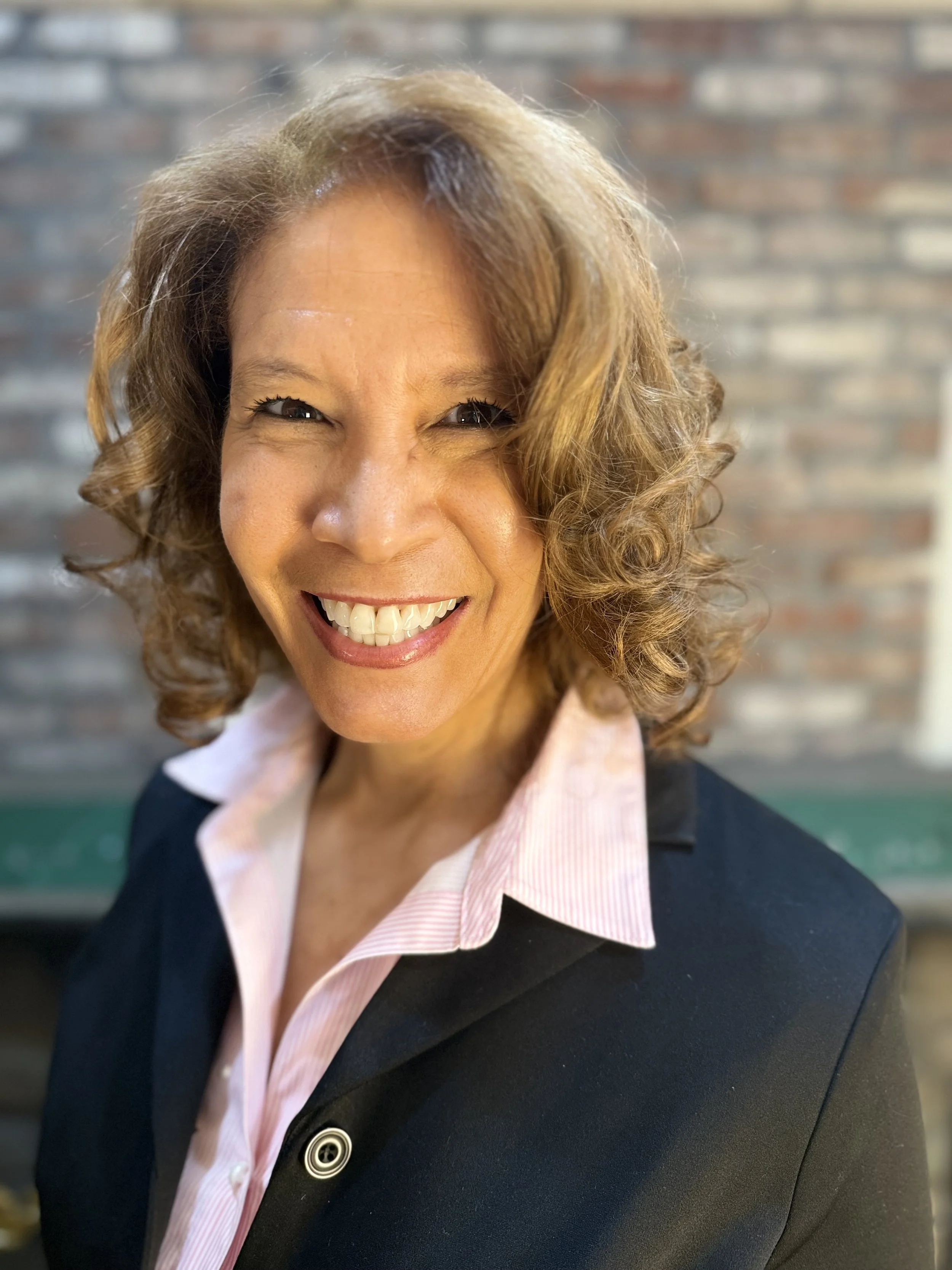 A woman with curly brown hair smiling at the camera, wearing a black blazer and white collared shirt, with a brick wall in the background.