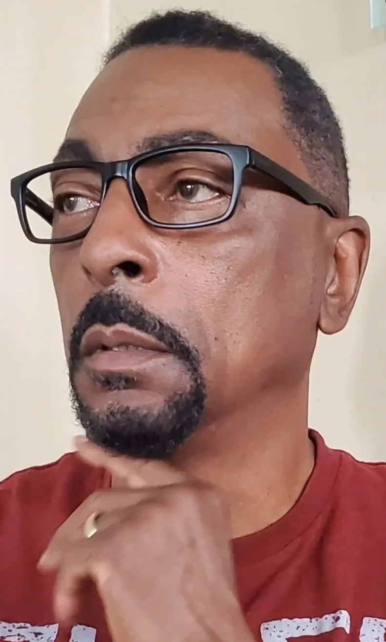 Close-up of a man with glasses, a beard, and a mustache, wearing a red shirt, touching his chin thoughtfully.