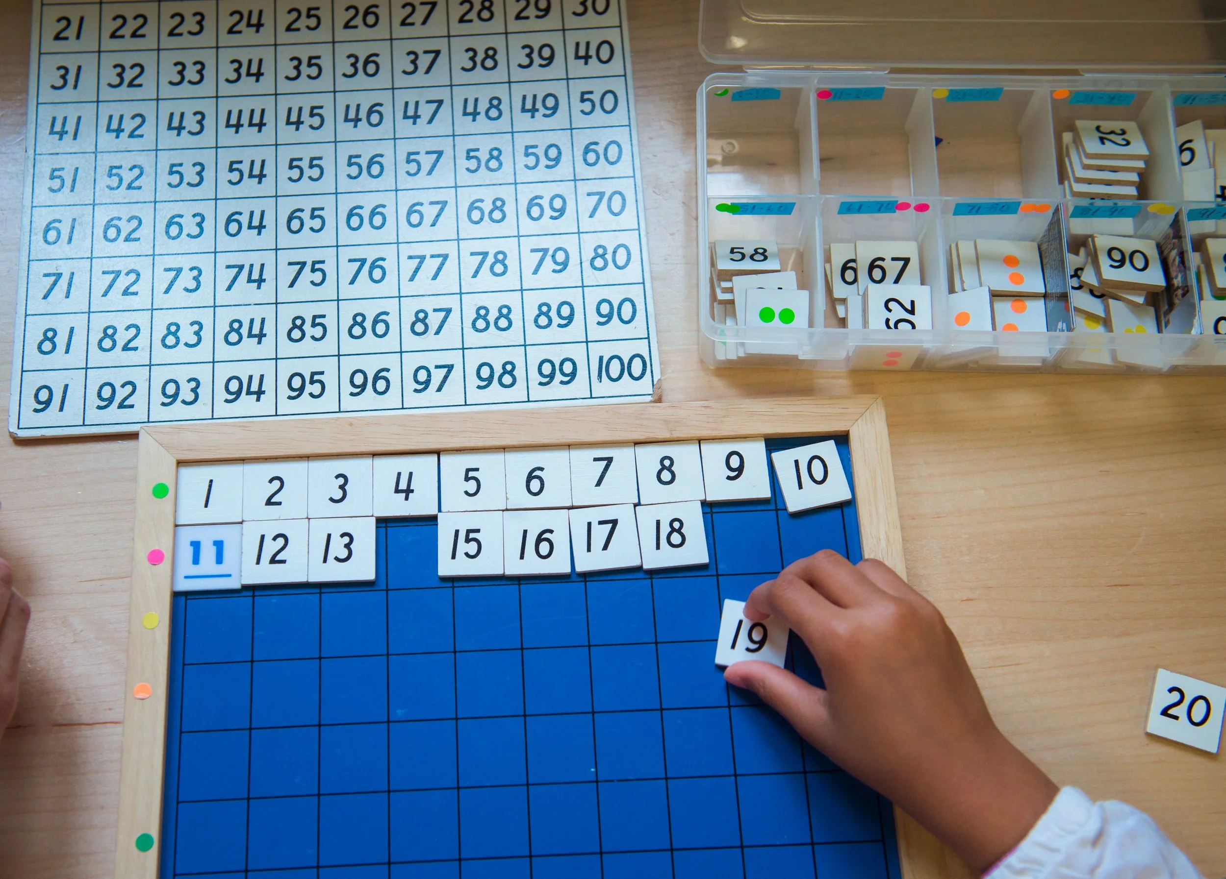 Child arranging number tiles on a blue grid board with a storage box and a number chart in the background.