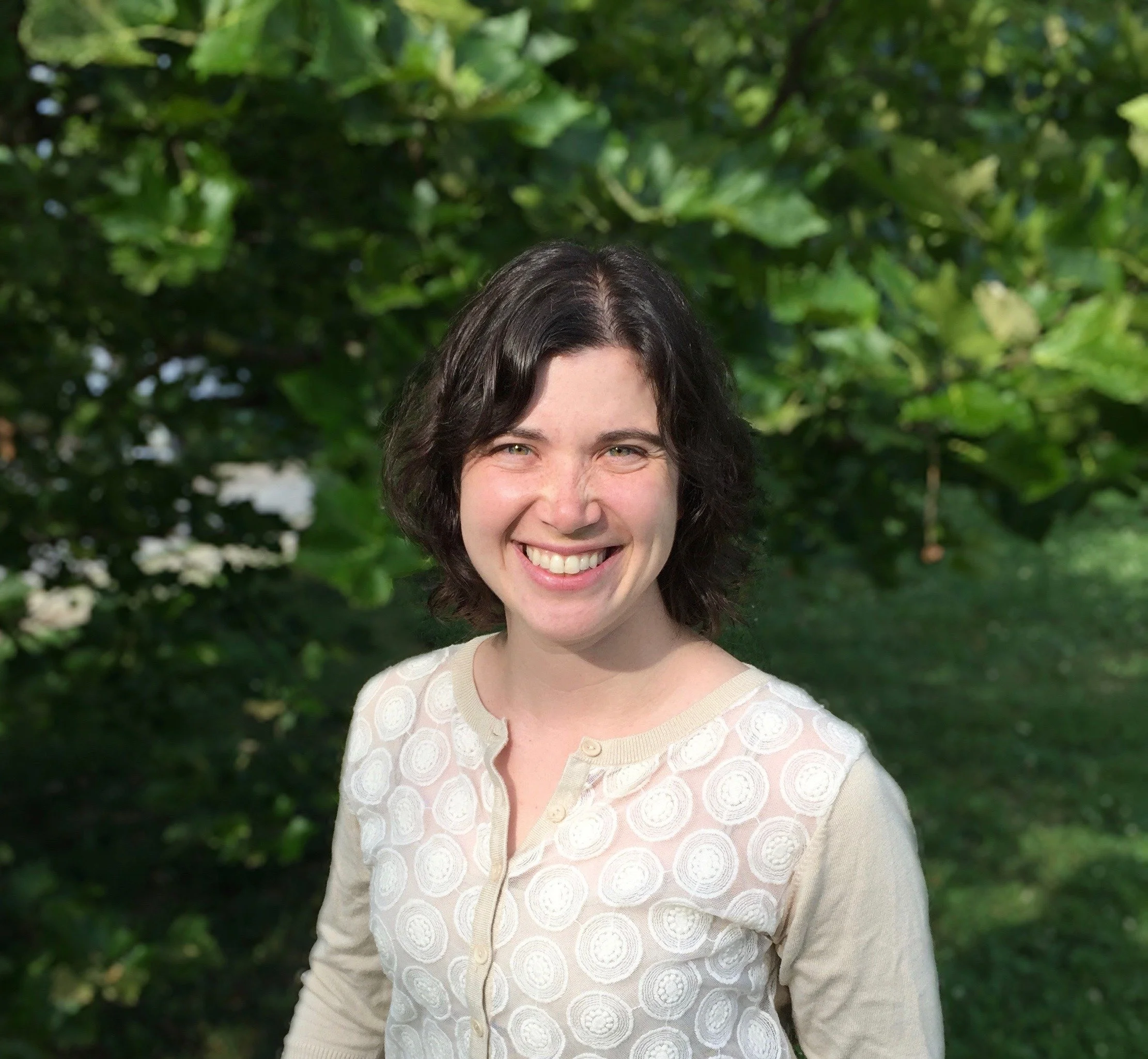 A woman with short, dark, wavy hair smiling outdoors with green trees and foliage in the background.