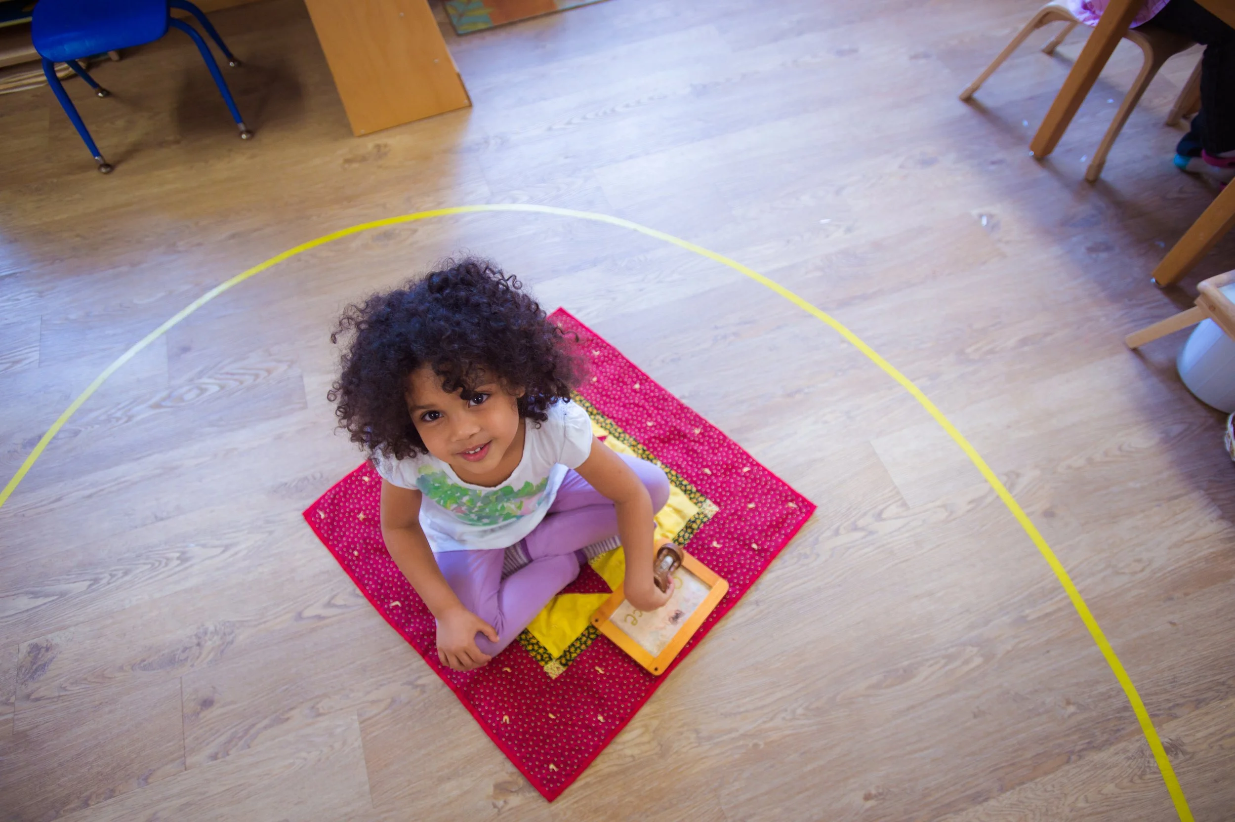 A young girl with curly hair sitting cross-legged on a red and yellow blanket on a wooden floor, looking up at the camera with a smile.