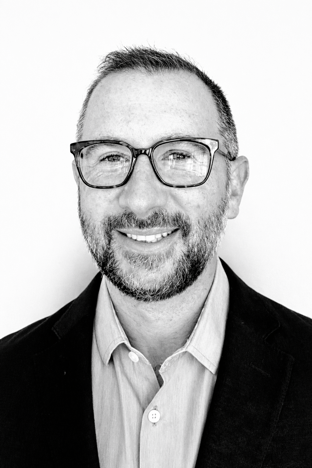 A man with curly dark hair and a beard, wearing a black suit, looking into camera with a slight smile. A white background