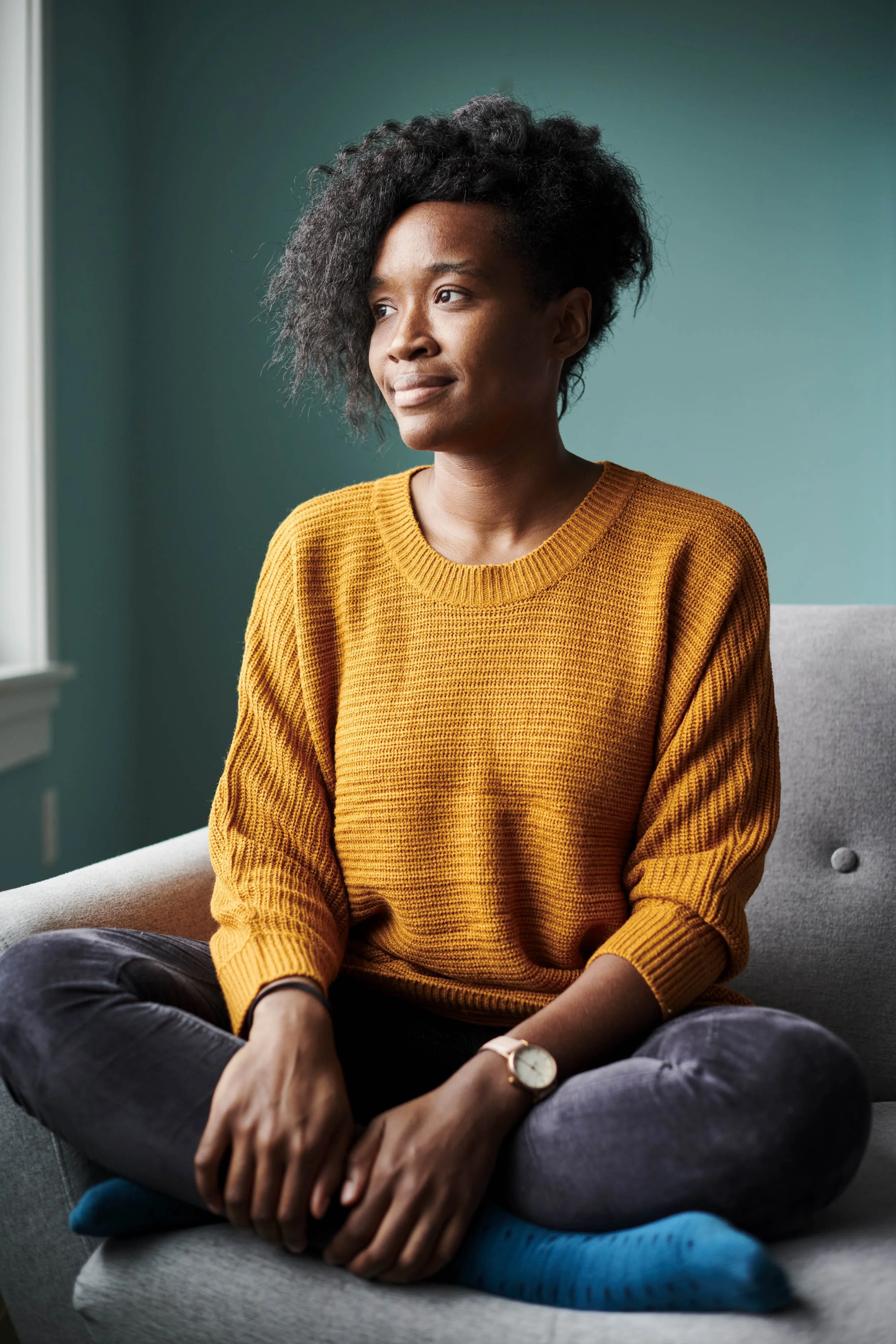 A woman with natural curly hair, sitting cross-legged on a gray sofa, wearing an orange sweater and blue socks, gazing out of a window with a thoughtful expression.