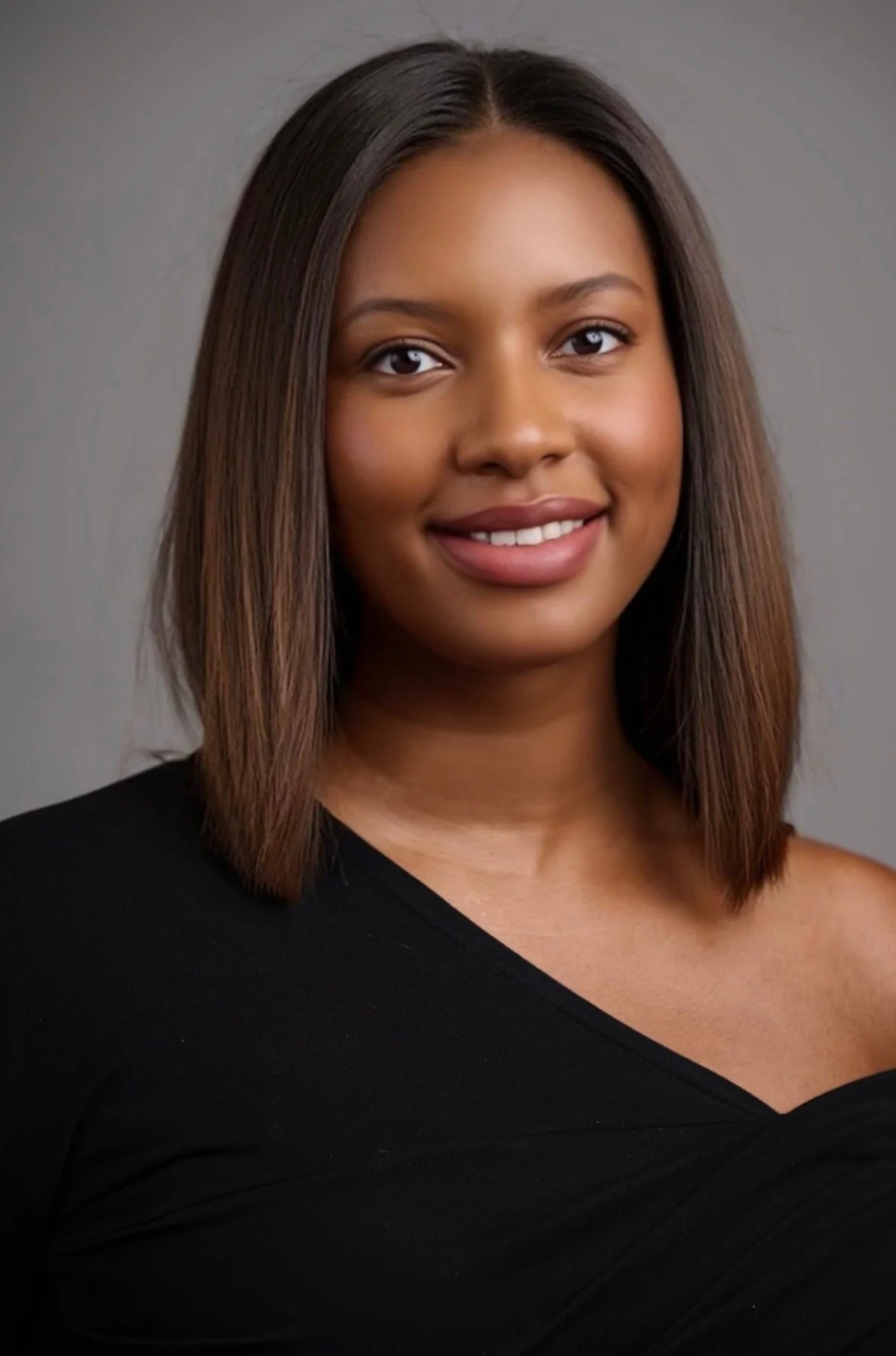 Portrait of a young woman with shoulder-length hair, smiling, wearing a black off-the-shoulder top against a gray background.