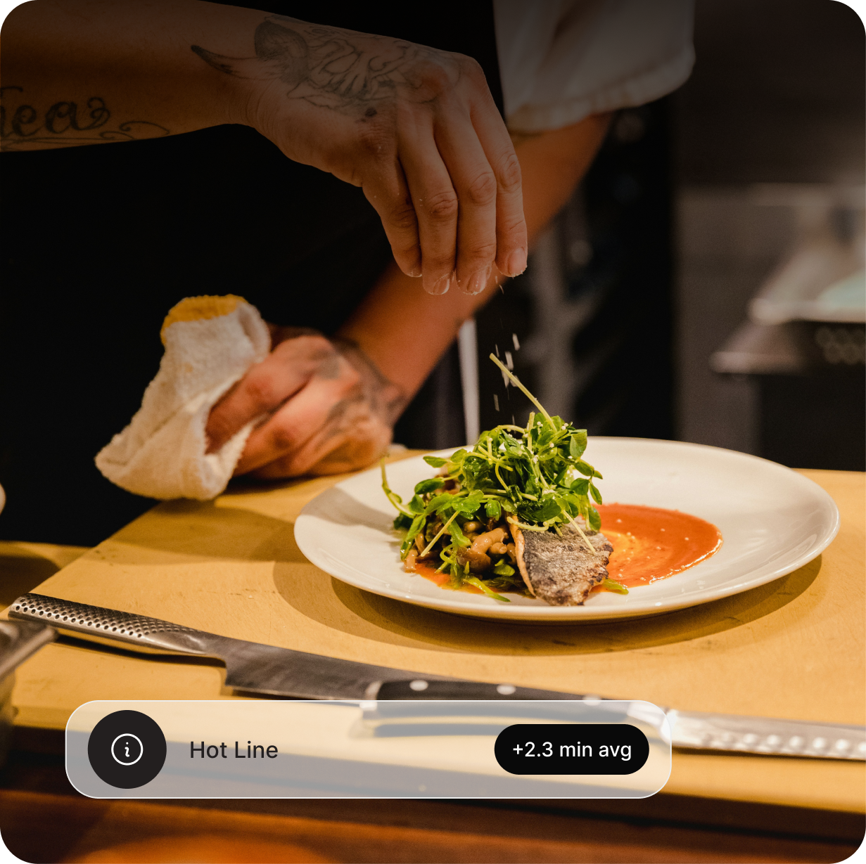 A chef garnishing a plated dish with microgreens, with a knife on the wooden cutting board in front.