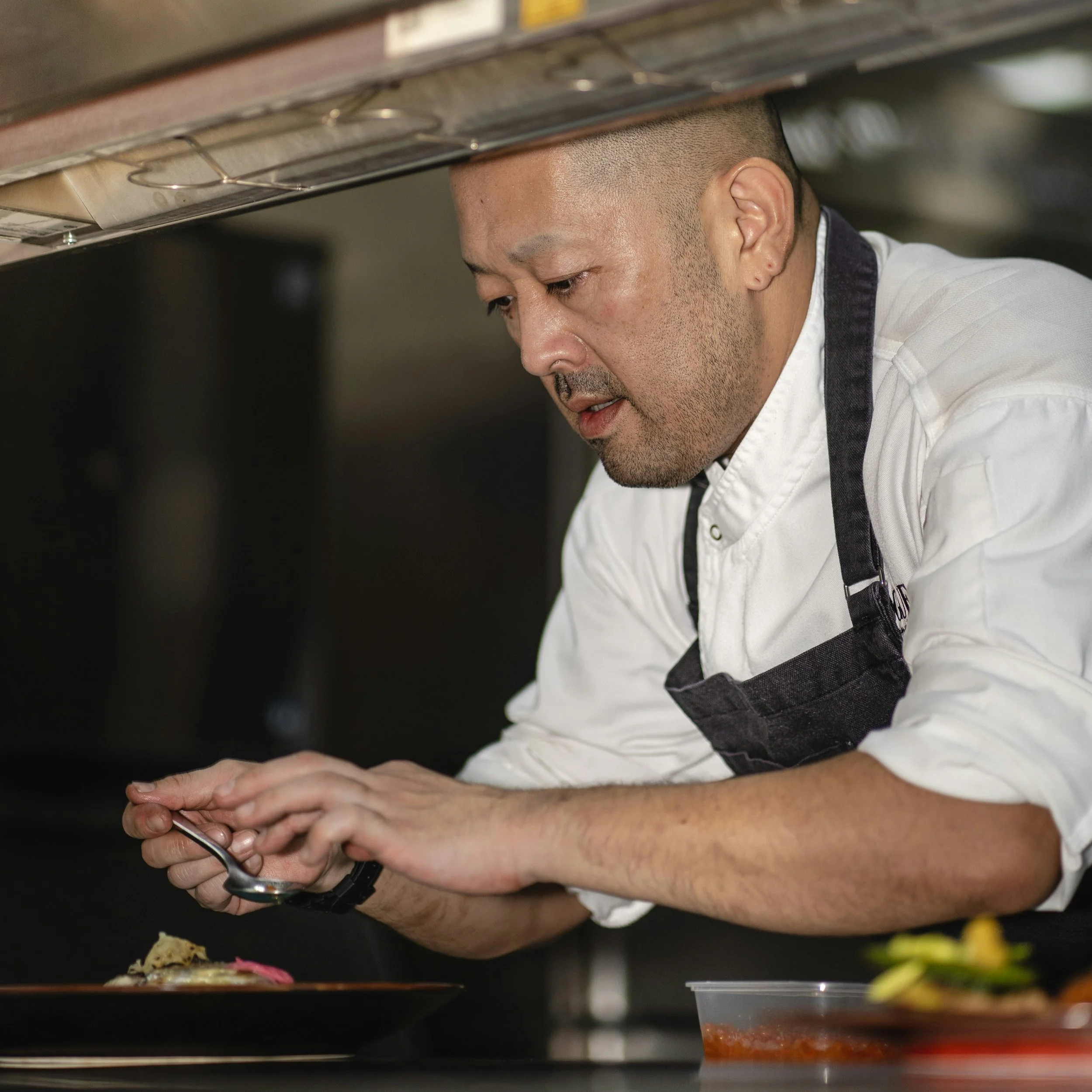 A chef in a white shirt and black apron garnishes a dish in a professional kitchen.