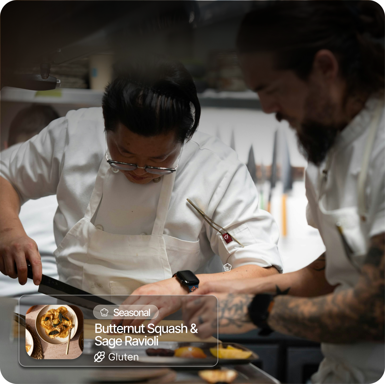 Two chefs in white uniforms working in a professional kitchen, preparing food with focused expressions. One chef wears glasses and a wristwatch, while the other has tattoos and a watch. A graphic overlay shows a dish named Butternut Squash & Sage Ravioli, which is seasonal and gluten-free.