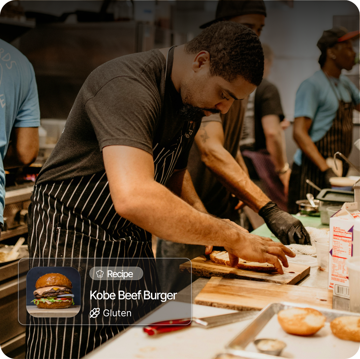 A man with a beard and black gloves making a Kobe beef burger in a kitchen, with other people in the background working.