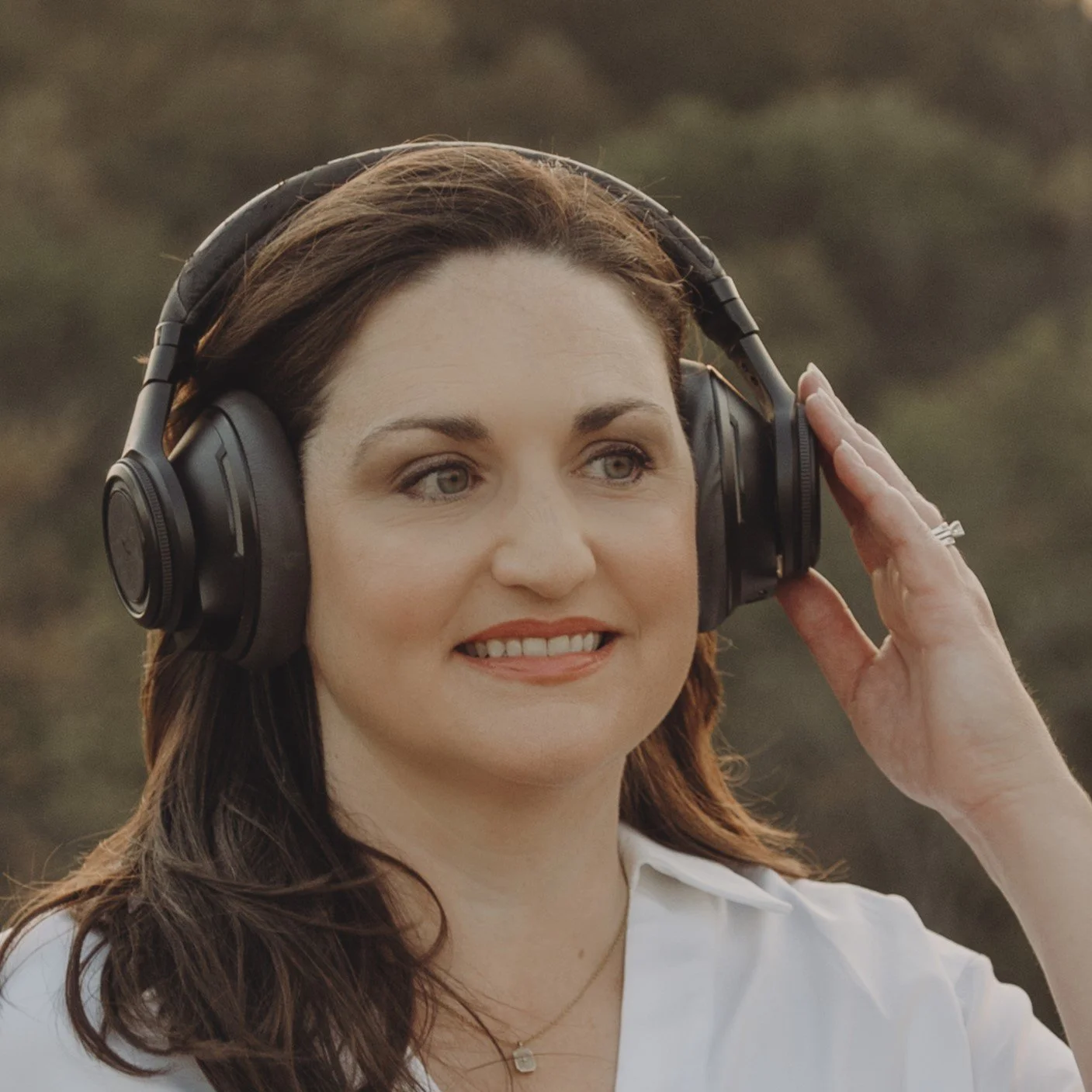 A woman with brown hear wearing black headphones looks off into the distance. Her left hand rests on the ear piece. She is smiling.