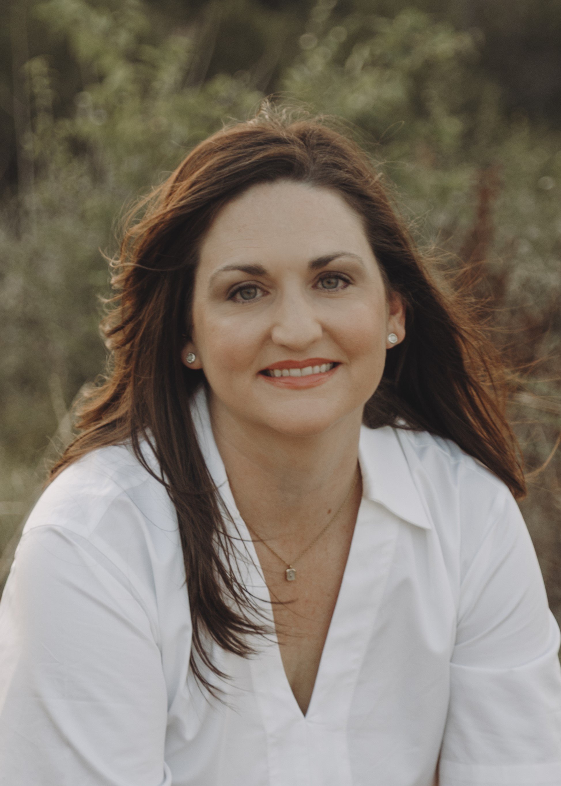 A woman smiles at the camera. She has wavy brown hair, green eyes, and white skin. She is wearing a white v-neck shirt and pearl earrings.