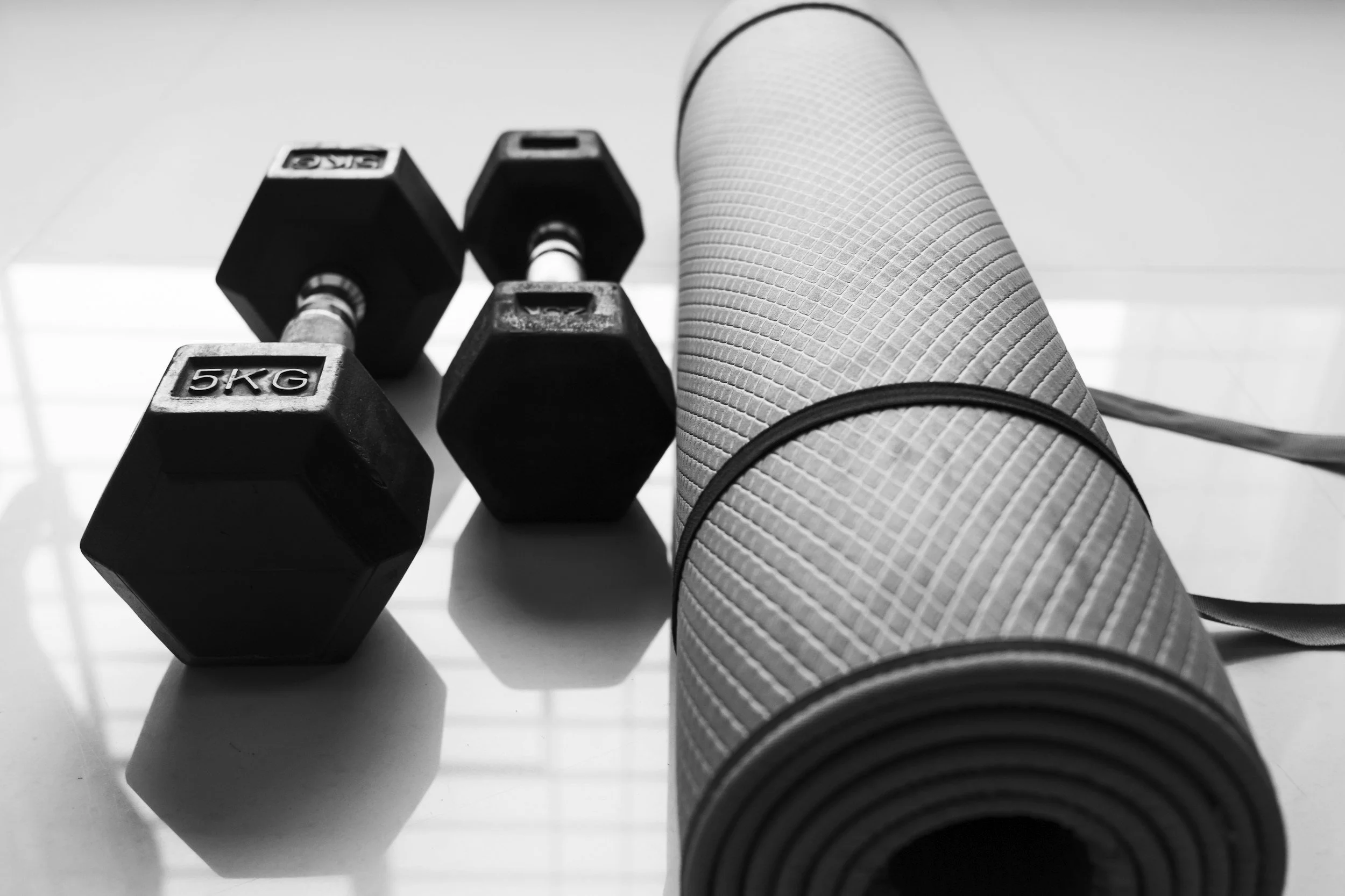 A rolled yoga mat and three dumbbells labeled 5kg on a reflective surface.