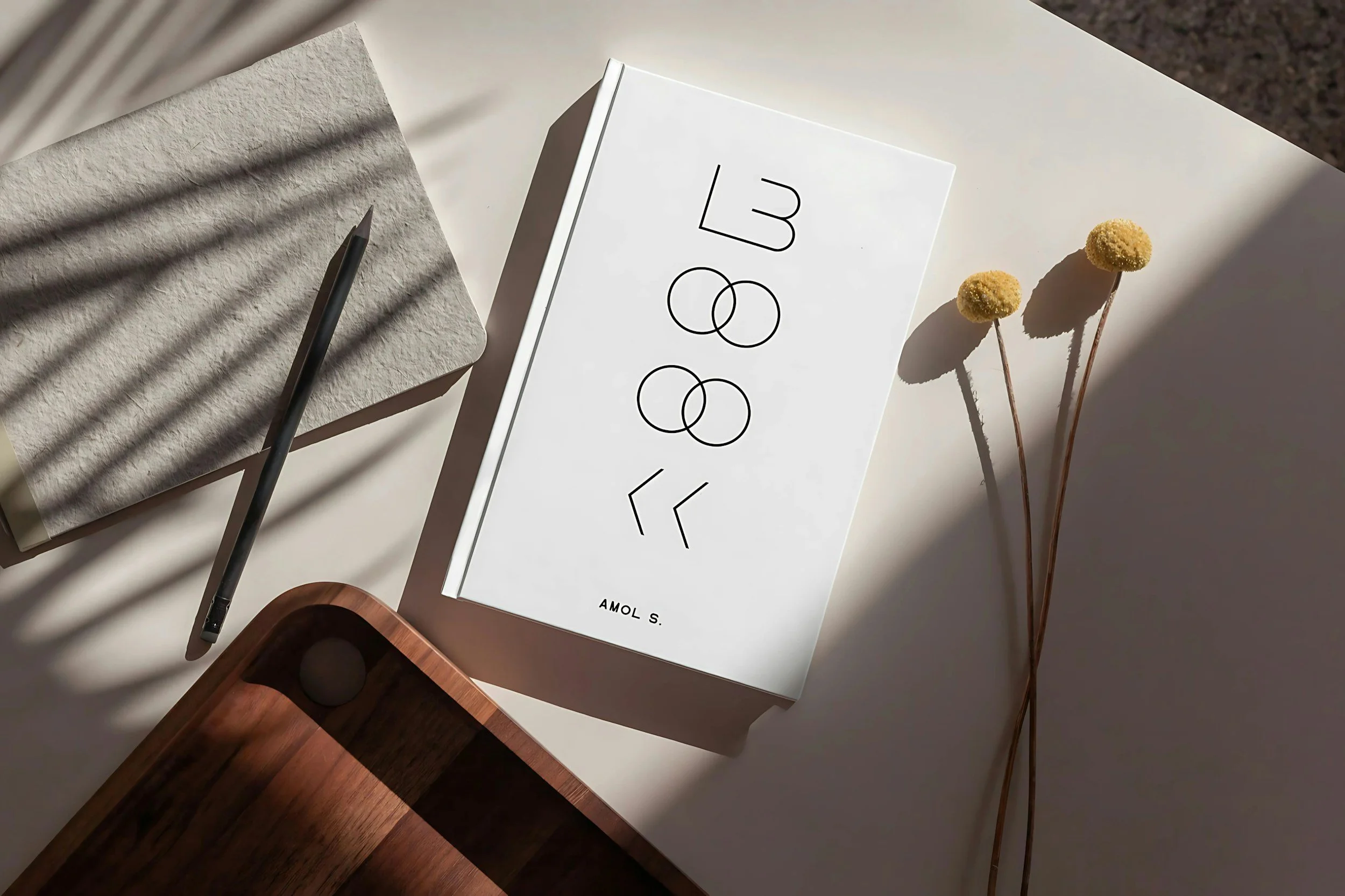 White book with black curved lines and the words 1988 on the cover, placed on a white surface beside a pencil, a closed textured notebook, dried yellow flowers, and a wooden object.