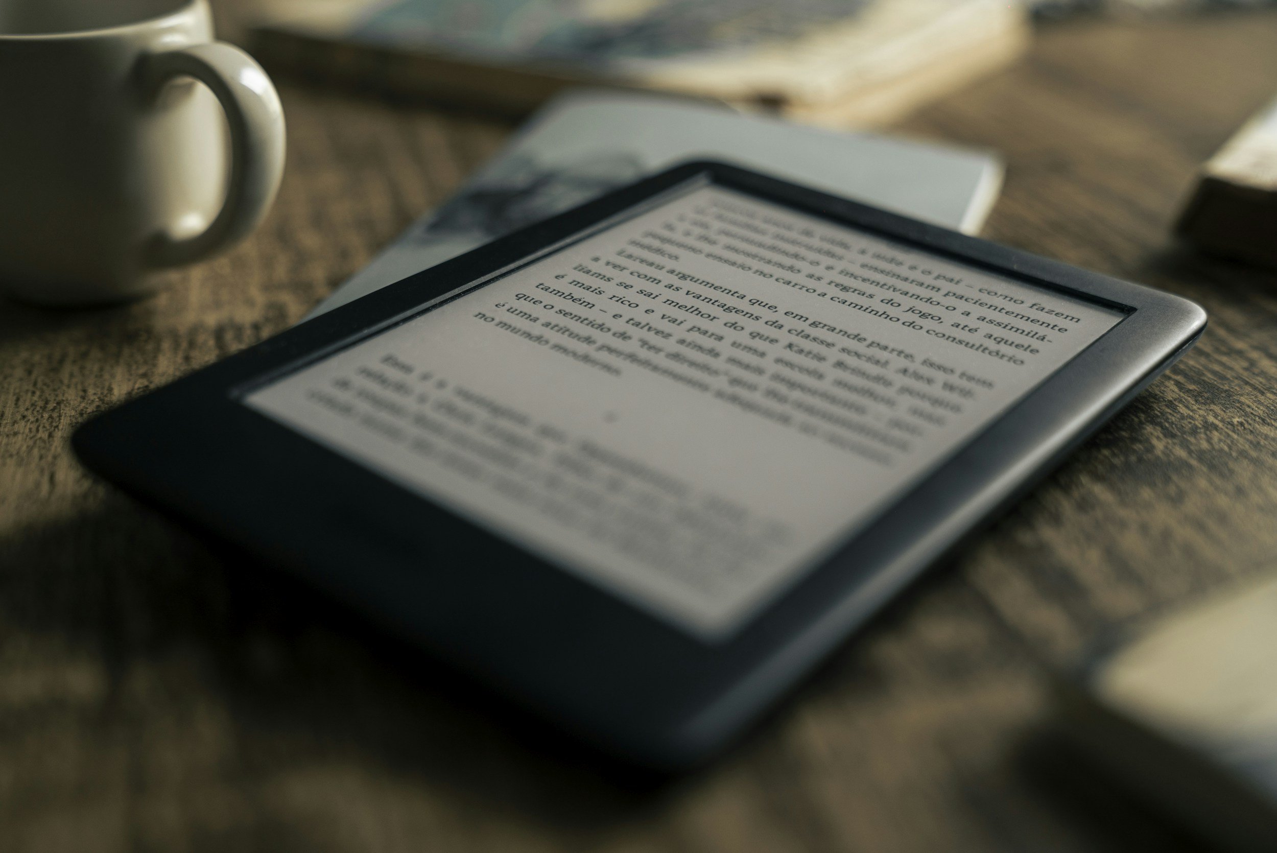 An e-reader device displaying text in Portuguese, placed on a wooden surface alongside a mug and books.