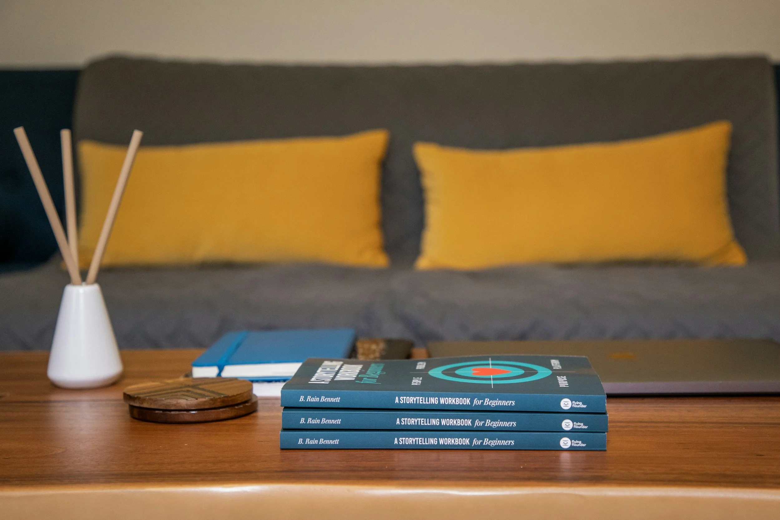 A wooden table with three blue books titled "A Storytelling Workbook for Beginners" by B. Rain Bennett stacked on top of each other, a closed silver laptop, a small wooden coaster, a white vase with reed diffuser sticks, a closed blue book, and a closed tablet; in the background, there is a sofa with dark gray fabric and two yellow cushions.