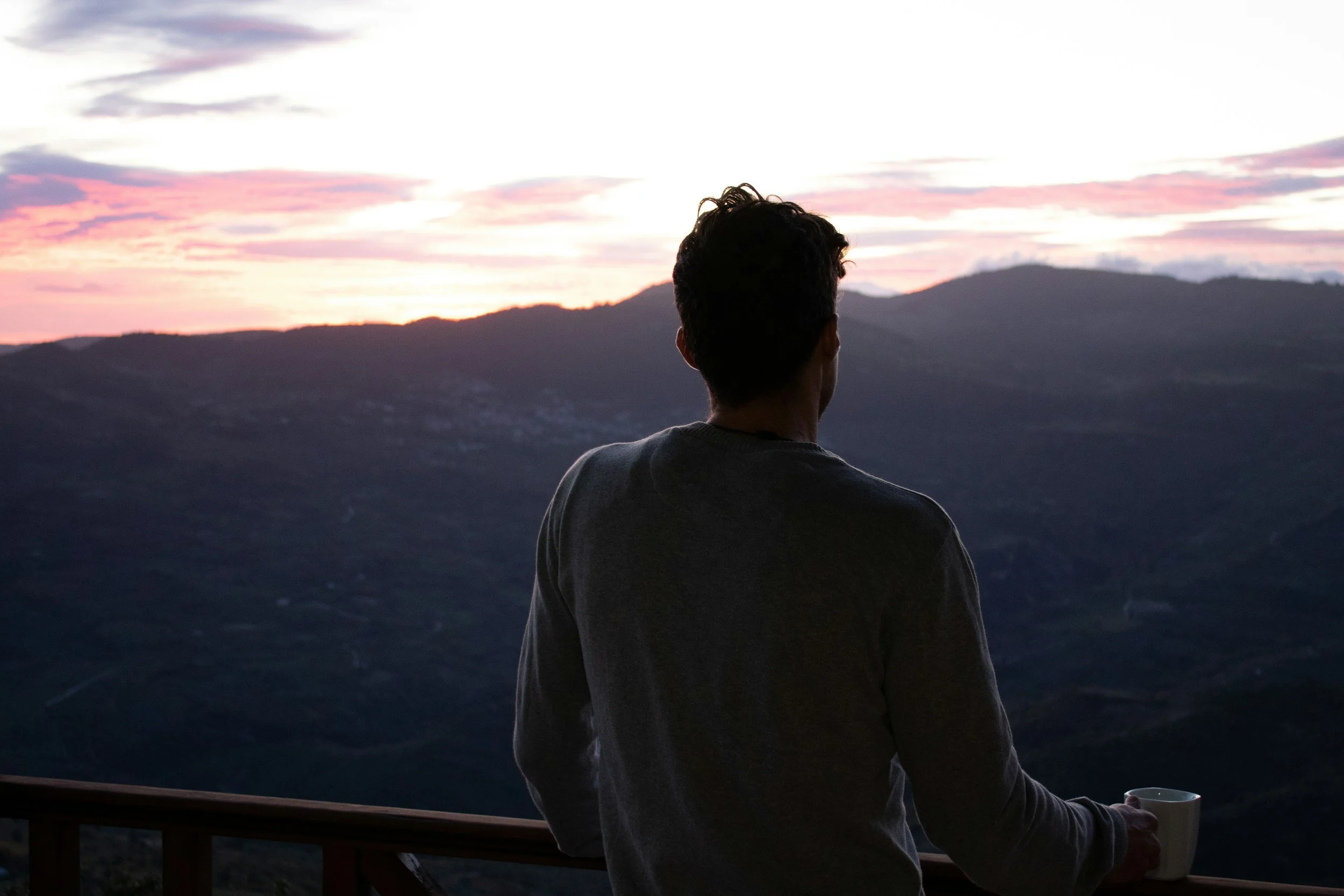 A person with dark, curly hair, wearing a gray long-sleeve shirt, standing on a balcony or deck, facing mountains and a sunset sky with clouds and pink hues, holding a white mug.