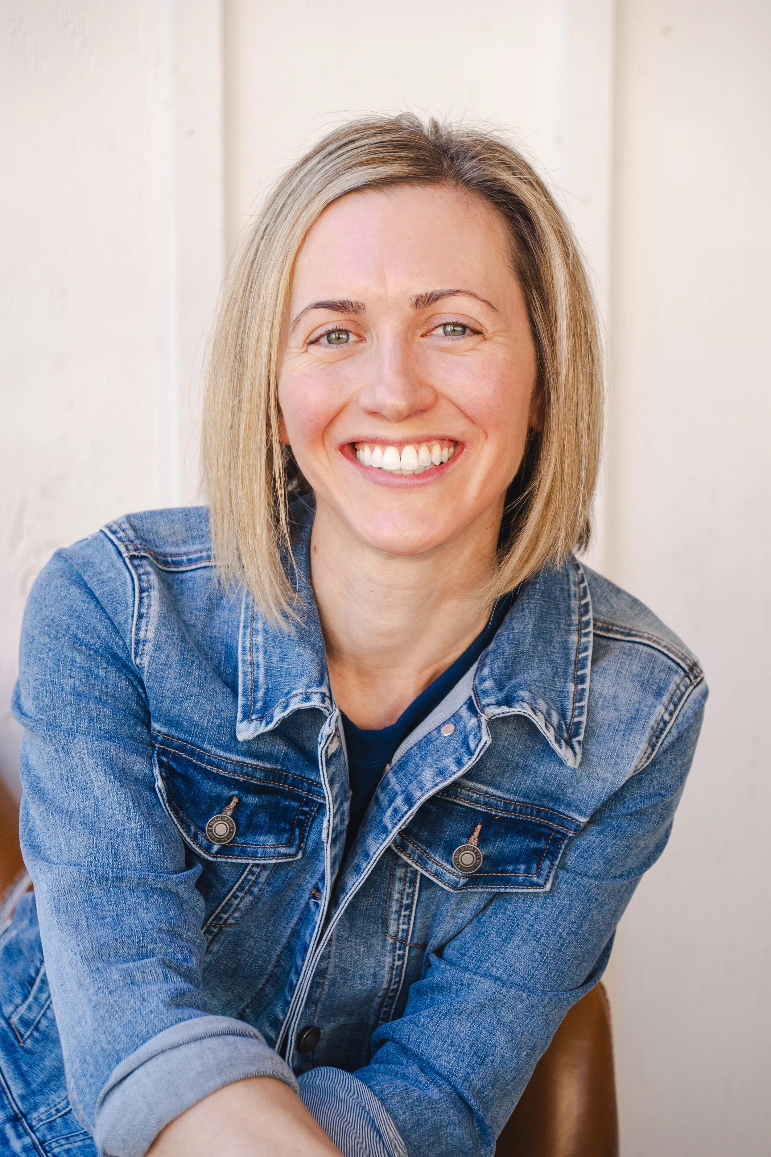 A woman with blonde hair smiling, wearing a denim jacket, sitting against a light-colored wall.