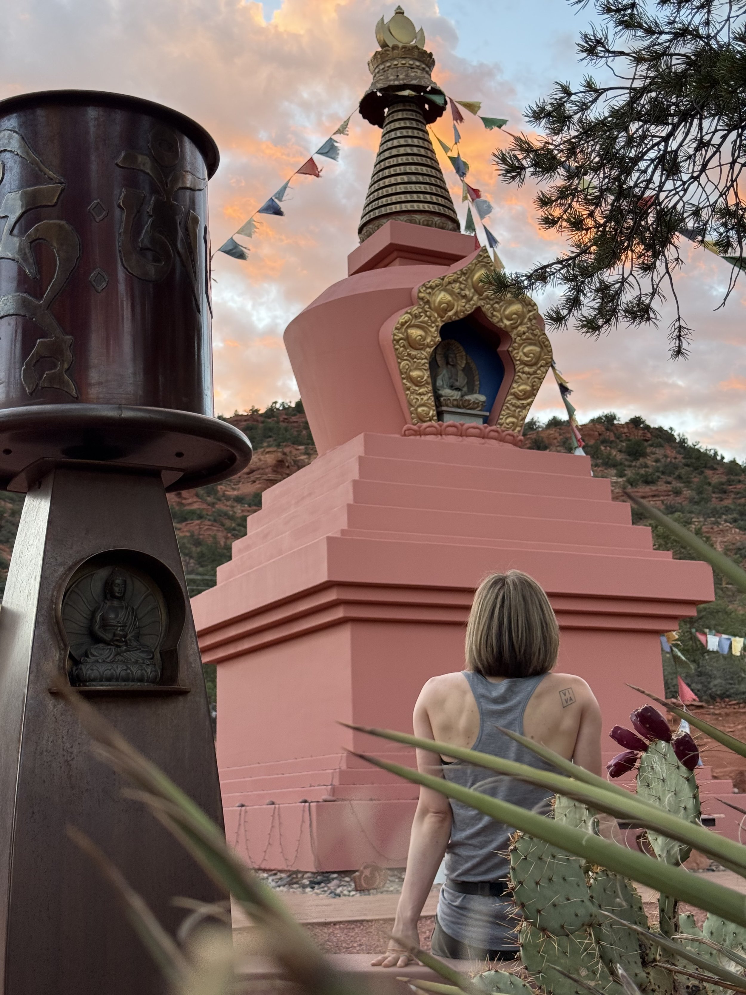 A woman sitting facing a pink Buddhist stupa with decorative gold accents and a small Buddha statue in an alcove, under a colorful sunset sky in Sedona.
