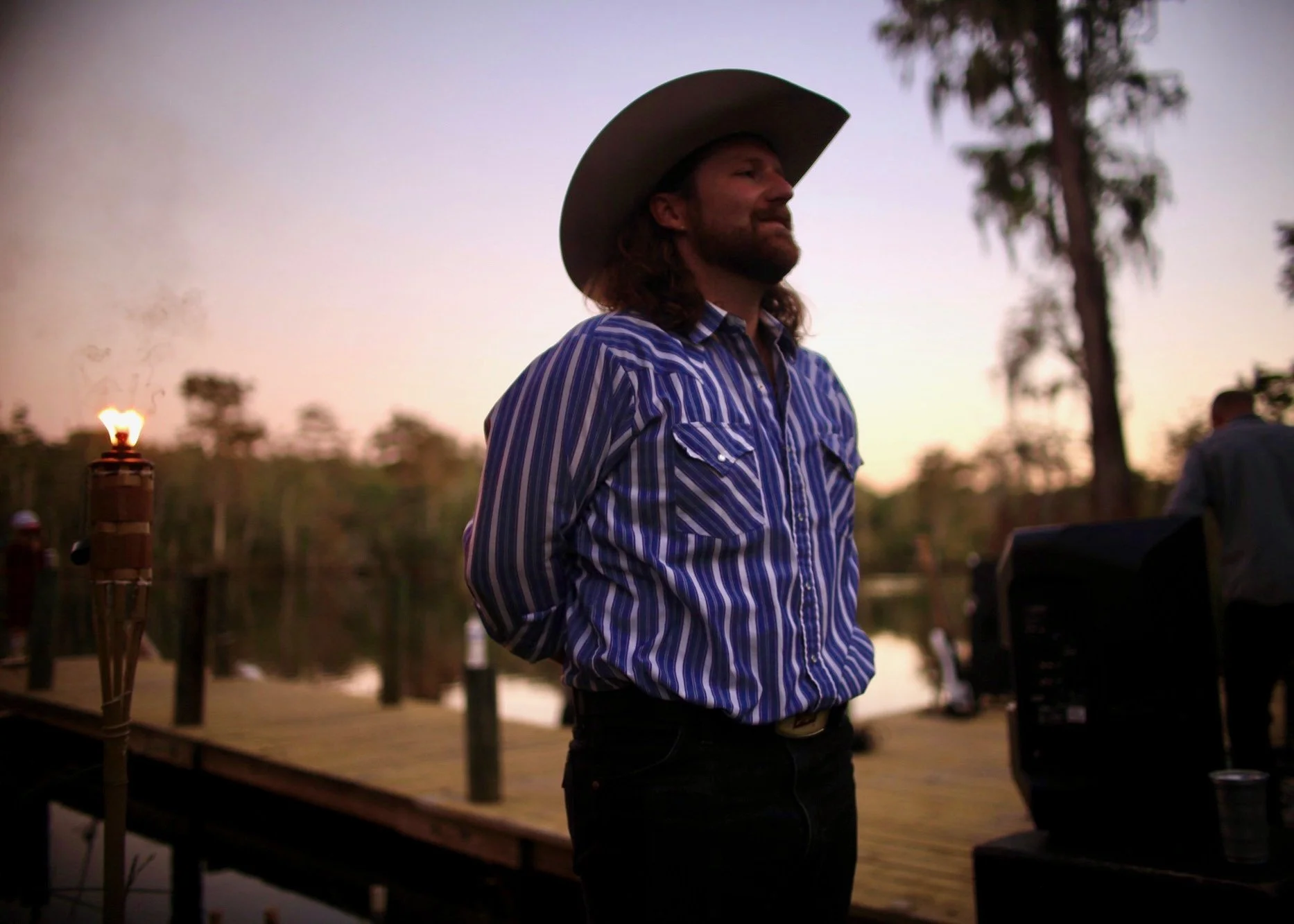 A man with long hair and a beard, wearing a cowboy hat and a blue striped shirt, stands outdoors during dusk near a lake and trees, with tiki torches and music equipment visible, smiling softly.