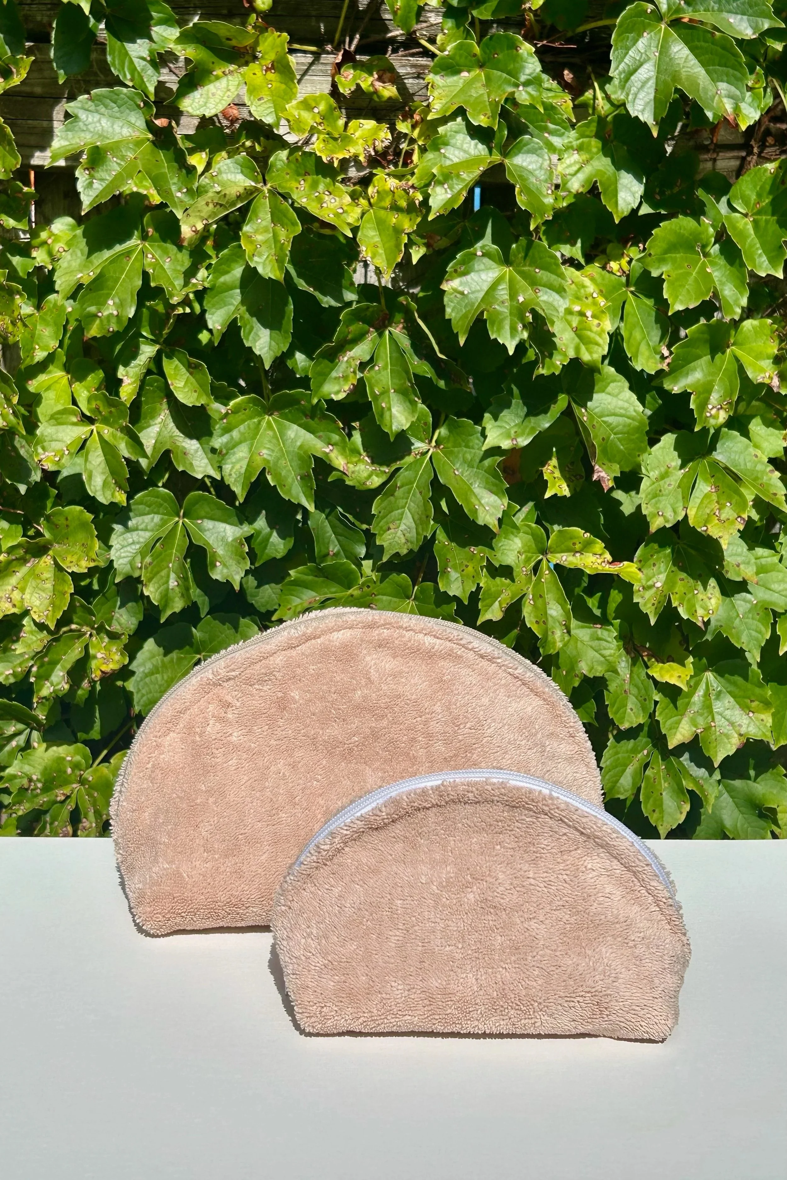 Two beige towels with zipper closures on a white surface, outdoors with green leafy plants in the background.