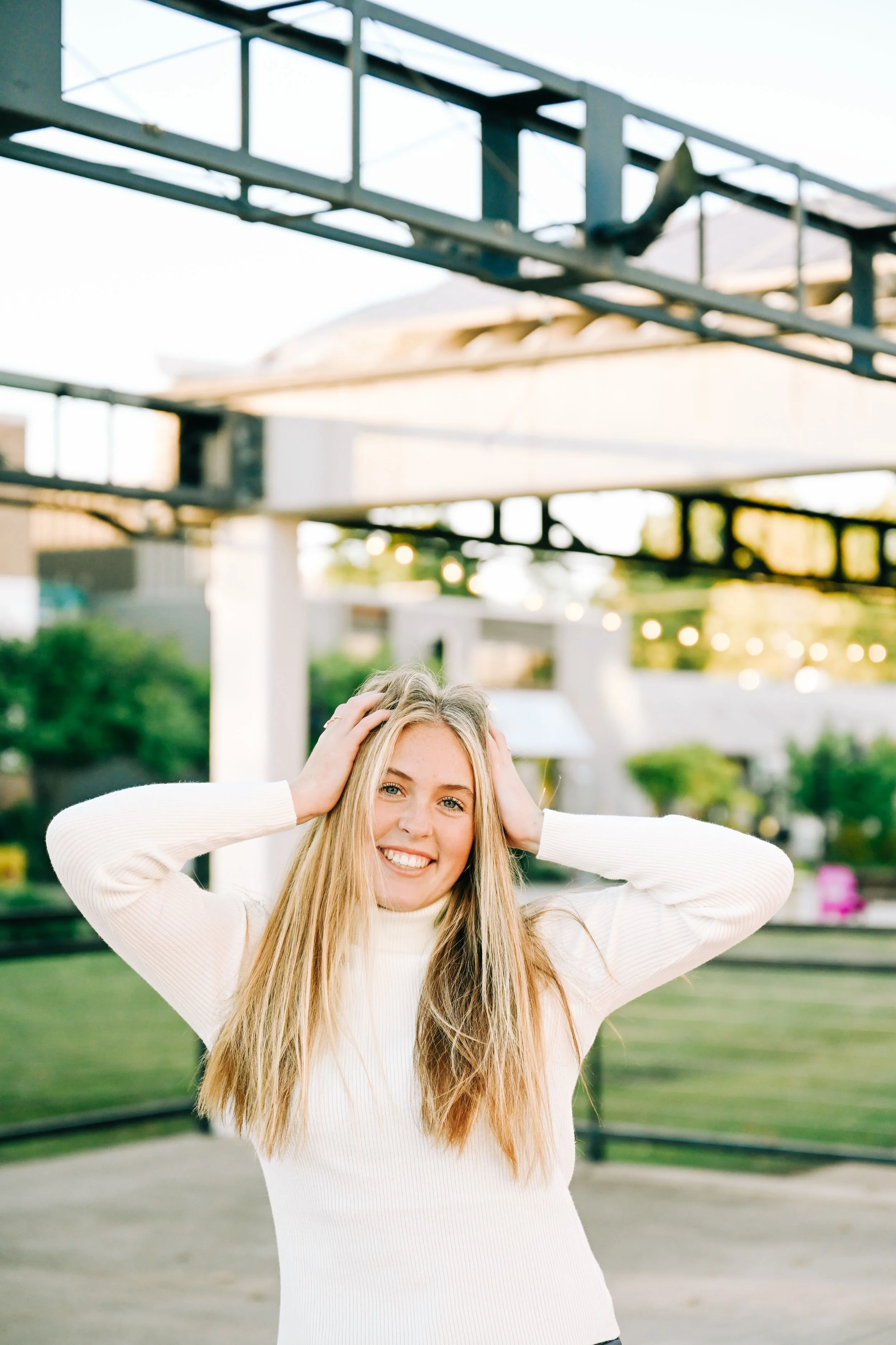 A woman with long blonde hair smiling and holding her hands on her head, standing outdoors in a park or garden area during daytime.