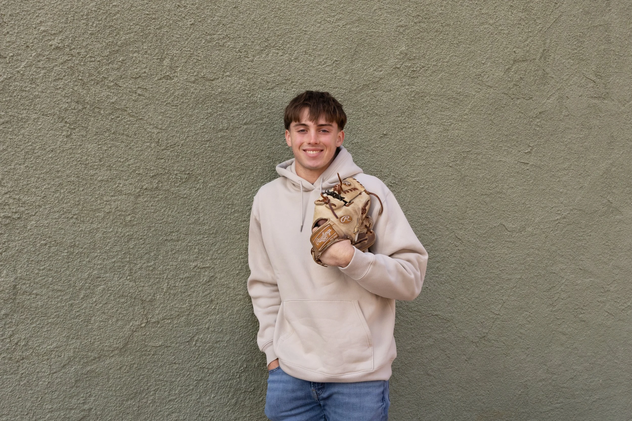 Young man in a beige hoodie holding a baseball glove, standing in front of a textured green wall.
