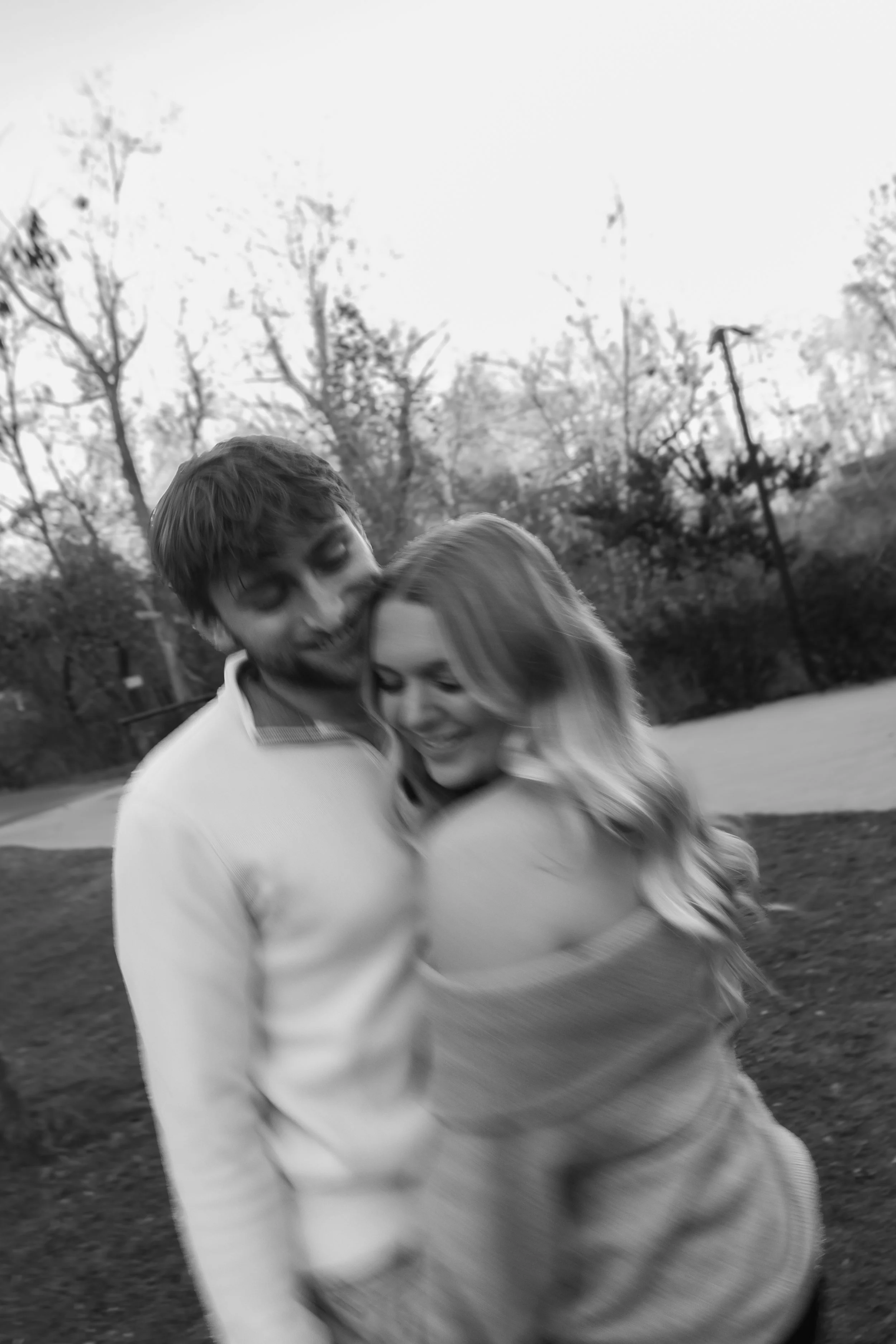 A black and white photo of a young man and woman hugging and smiling outdoors, with trees and a pathway in the background.