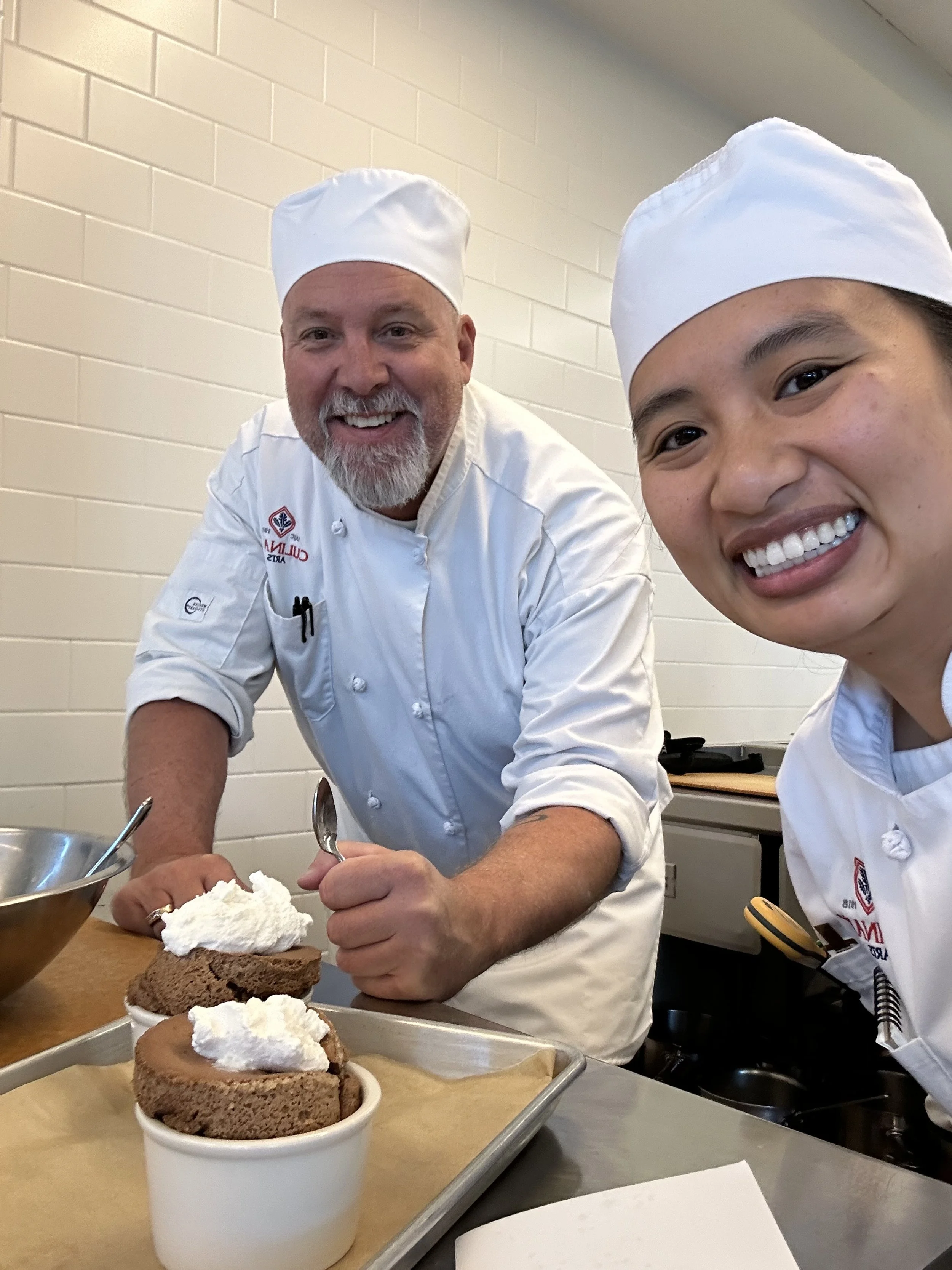 Two chefs, a man and a woman, wearing white chef uniforms and hats, smiling at the camera while preparing desserts in a kitchen.