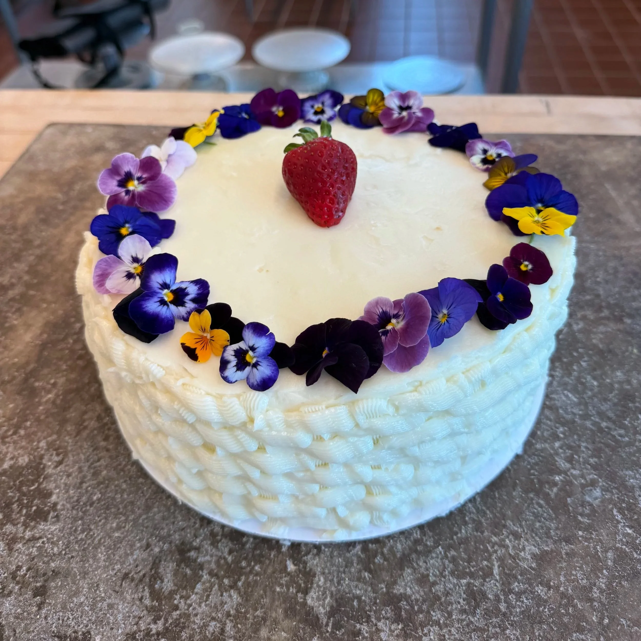 White cake decorated with a ring of purple, yellow, and violet edible flowers and a single strawberry on top, placed on a gray surface.